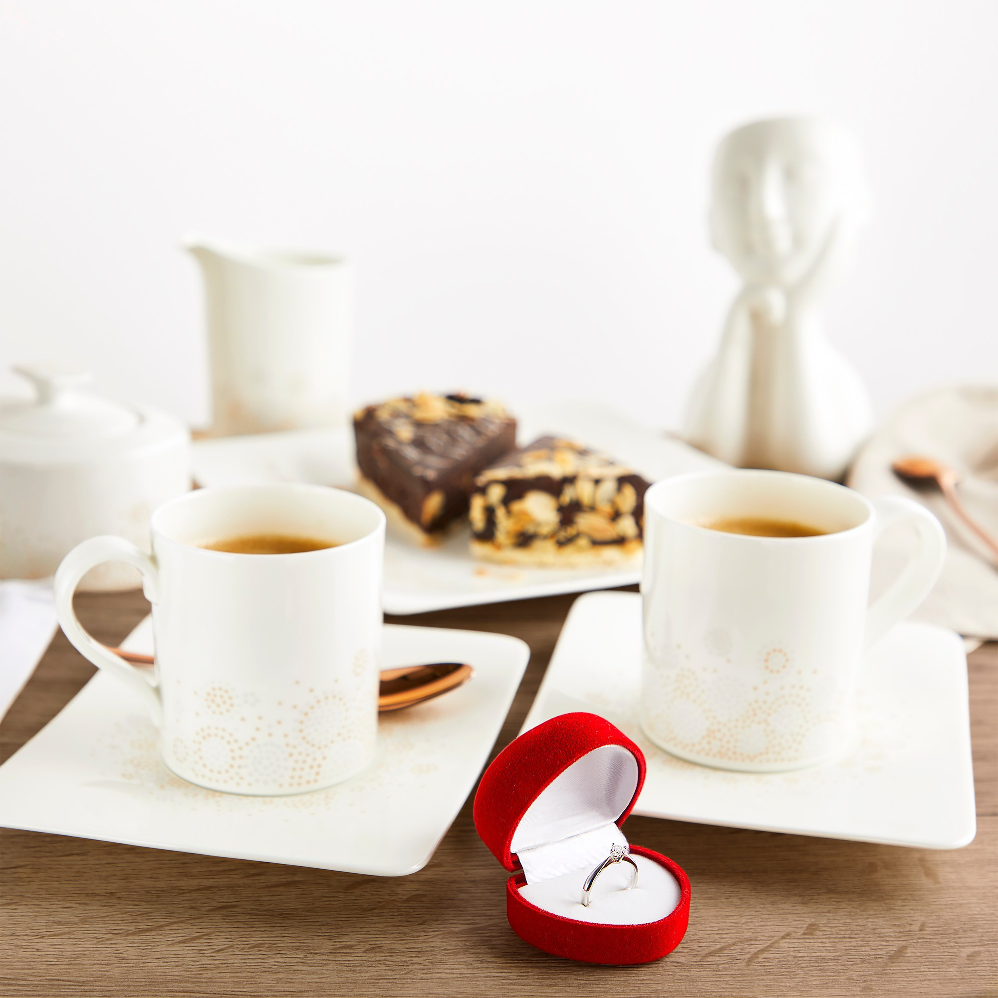 Photographer Roman Djuzev - Still life with a ring for a marriage proposal. In the foreground is a ring in a box, two cups of coffee, in the background a cute ceramic figurine and cakes.