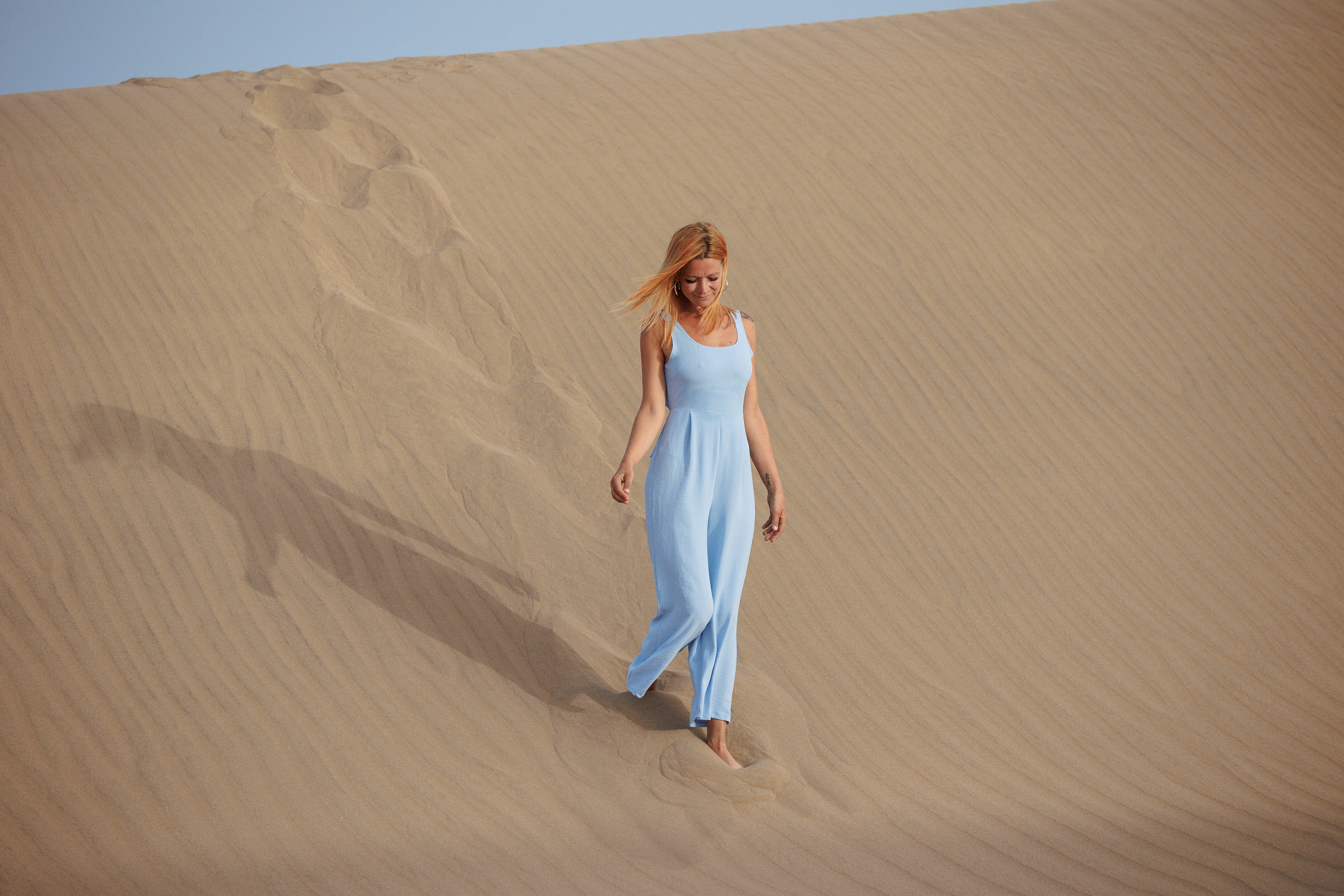 Immerse yourself in the breathtaking sight of a woman adorned in a captivating blue dress, gracefully traversing the sand dunes of Maspalomas for an unforgettable portrait photo shoot.