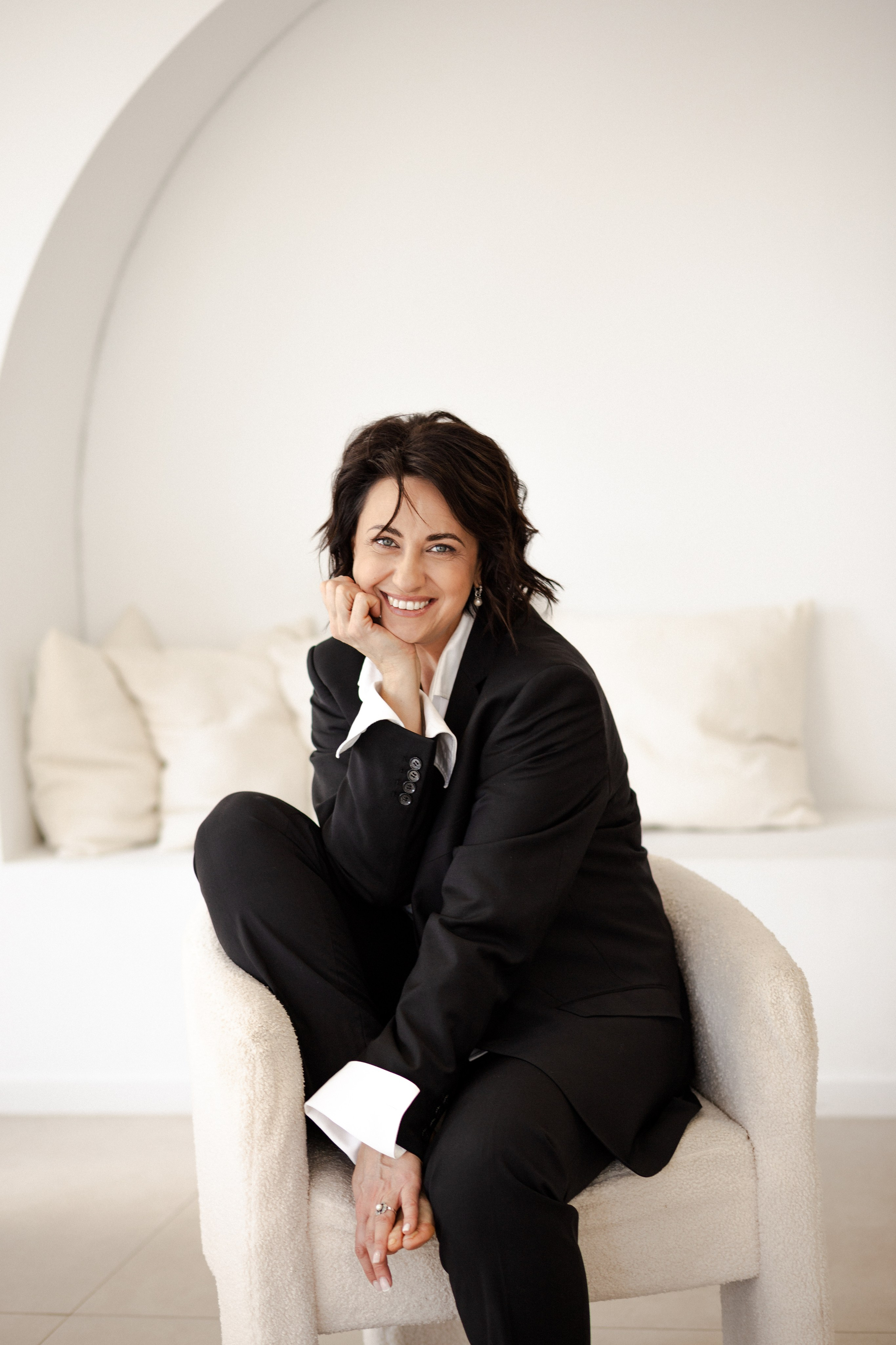 Профессиональный портрет улыбающейся женщины в черном костюме, удобно сидящей в современном белом кресле на фоне минималистичного интерьера в фотостудии в Валенсии, Испания