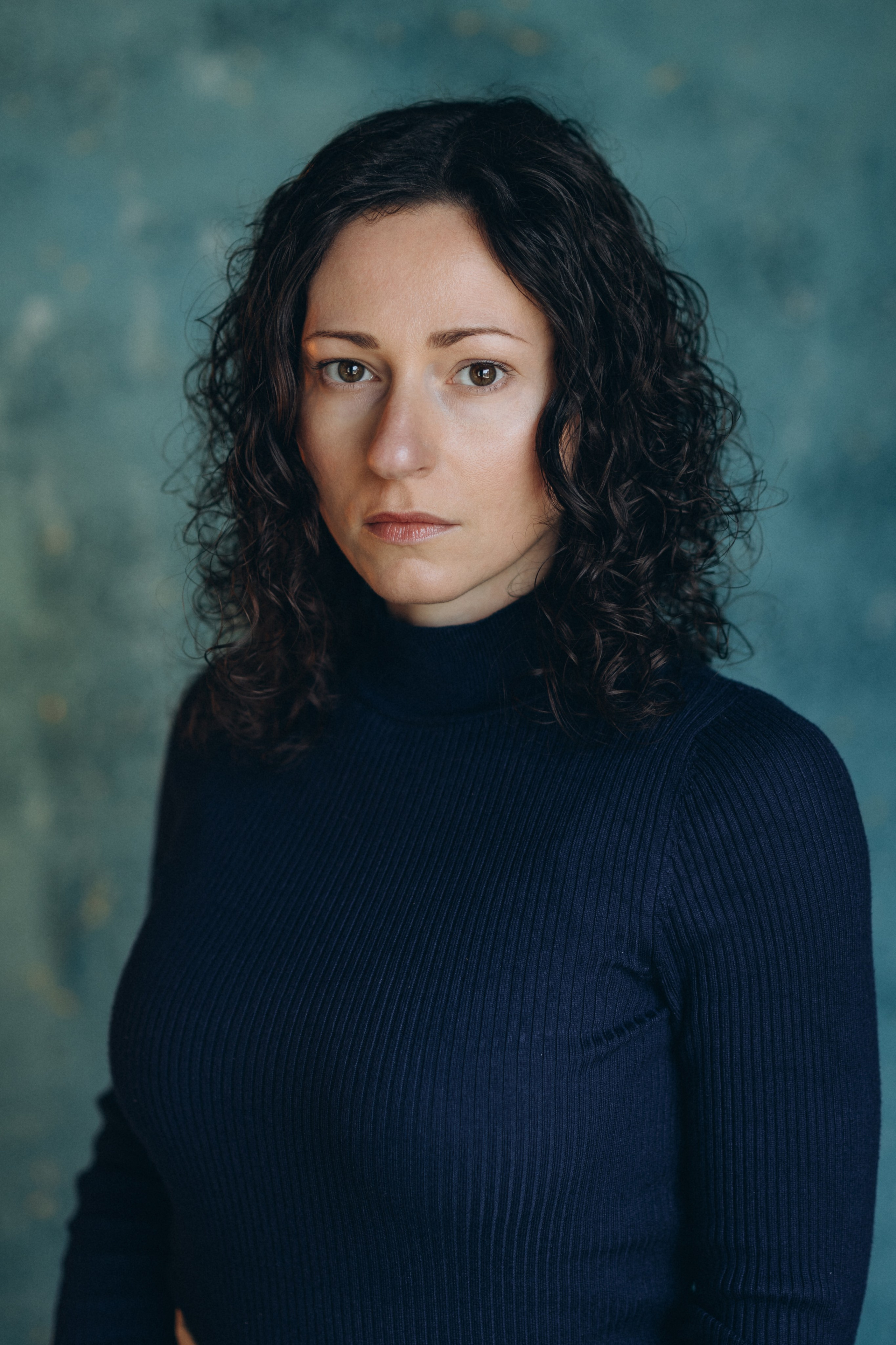 Actor headshot of a serious woman with curly dark hair in a navy blue top, standing against a textured blue background