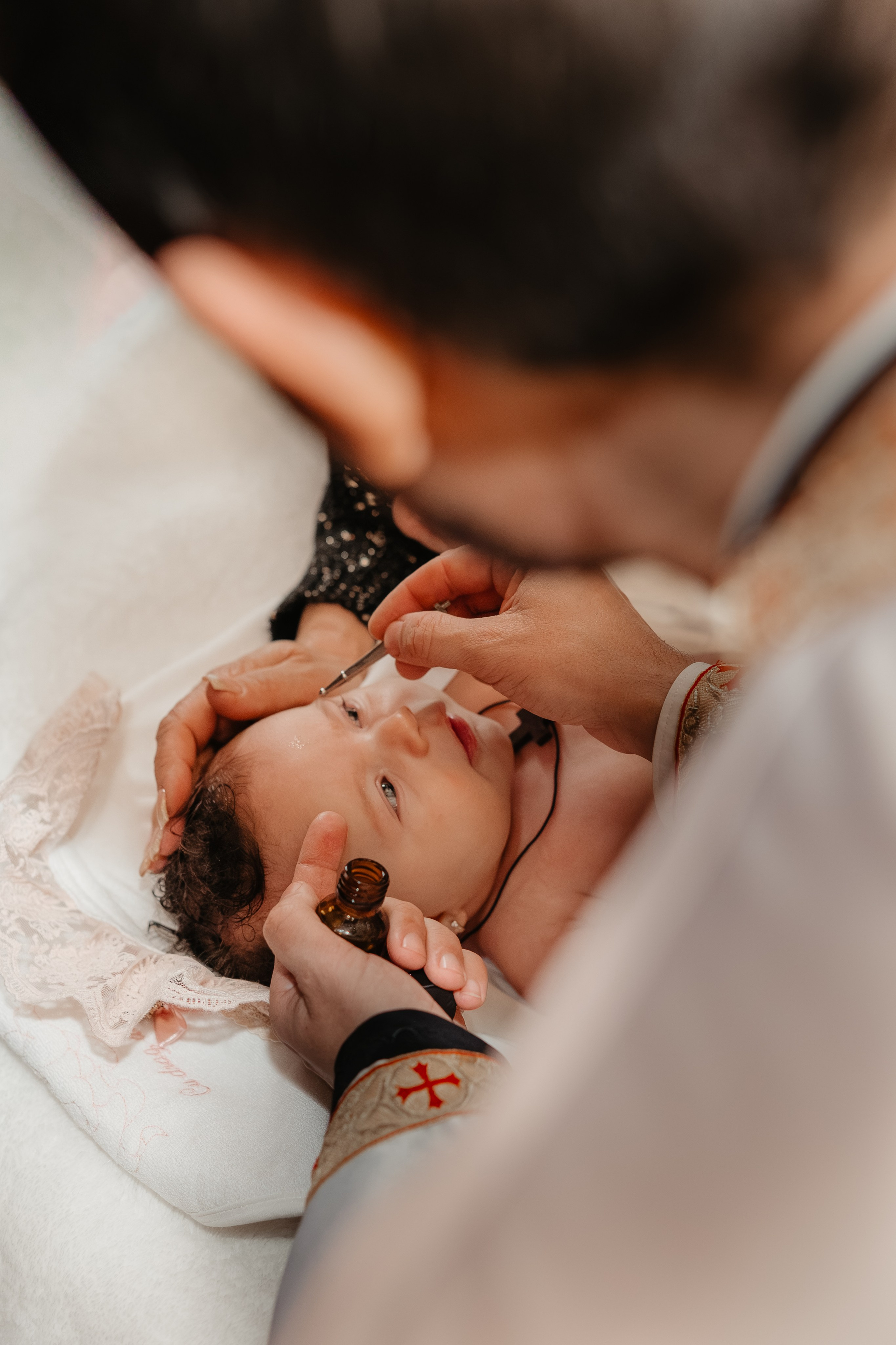 A doua ungere cu mir sfânt a pruncului Eveline Maria, pe masa botezului. Preotul aplică mirul cu grijă pe frunte, încheind ritualul solemn. Fotografie intimă a unui moment final și profund simbolic.