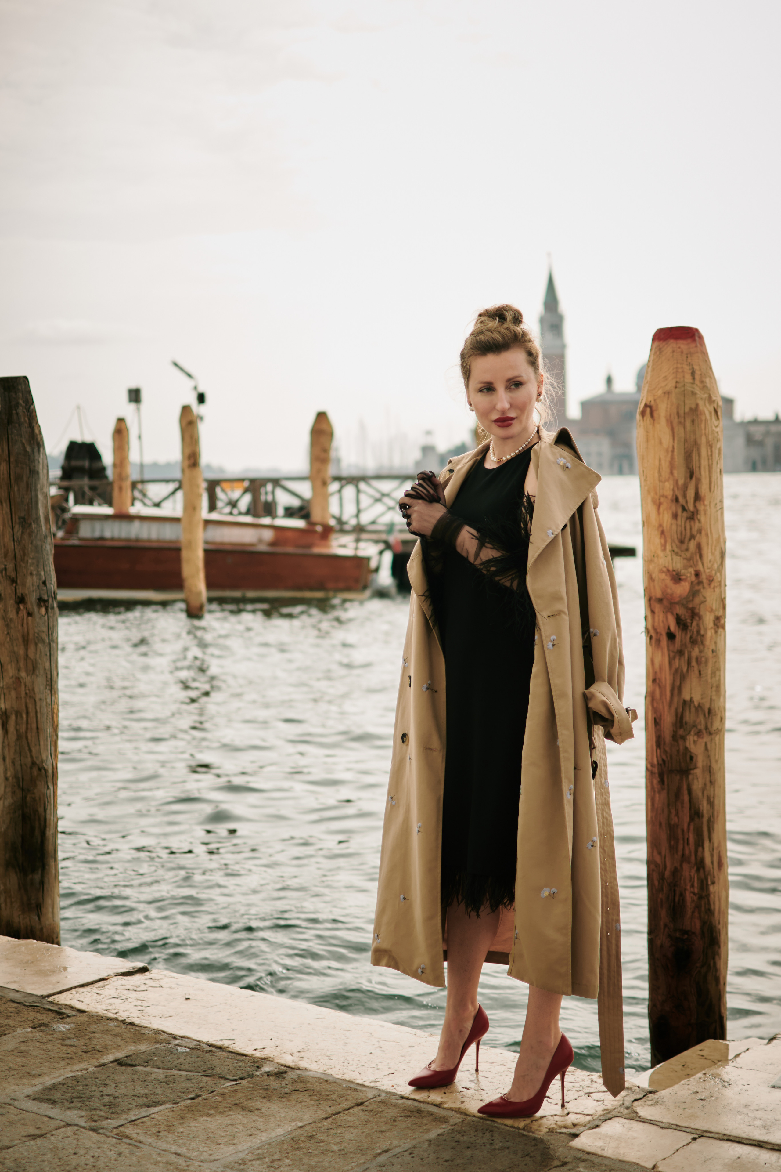 A young woman with a bright smile stands on San Marco square in Venice. She wears a flowing yellow sundress that complements the warm sunlight reflecting off the water behind her. The red and white striped poles of a gondola rest in the background, adding to the Italian charm of the scene. She looks relaxed and carefree, enjoying the beautiful day in this timeless city. Fashion blogger shoot Venice. Portrait and Fashion Photographer in Venice, Italy. Instagram Photos with lifestyle photographer