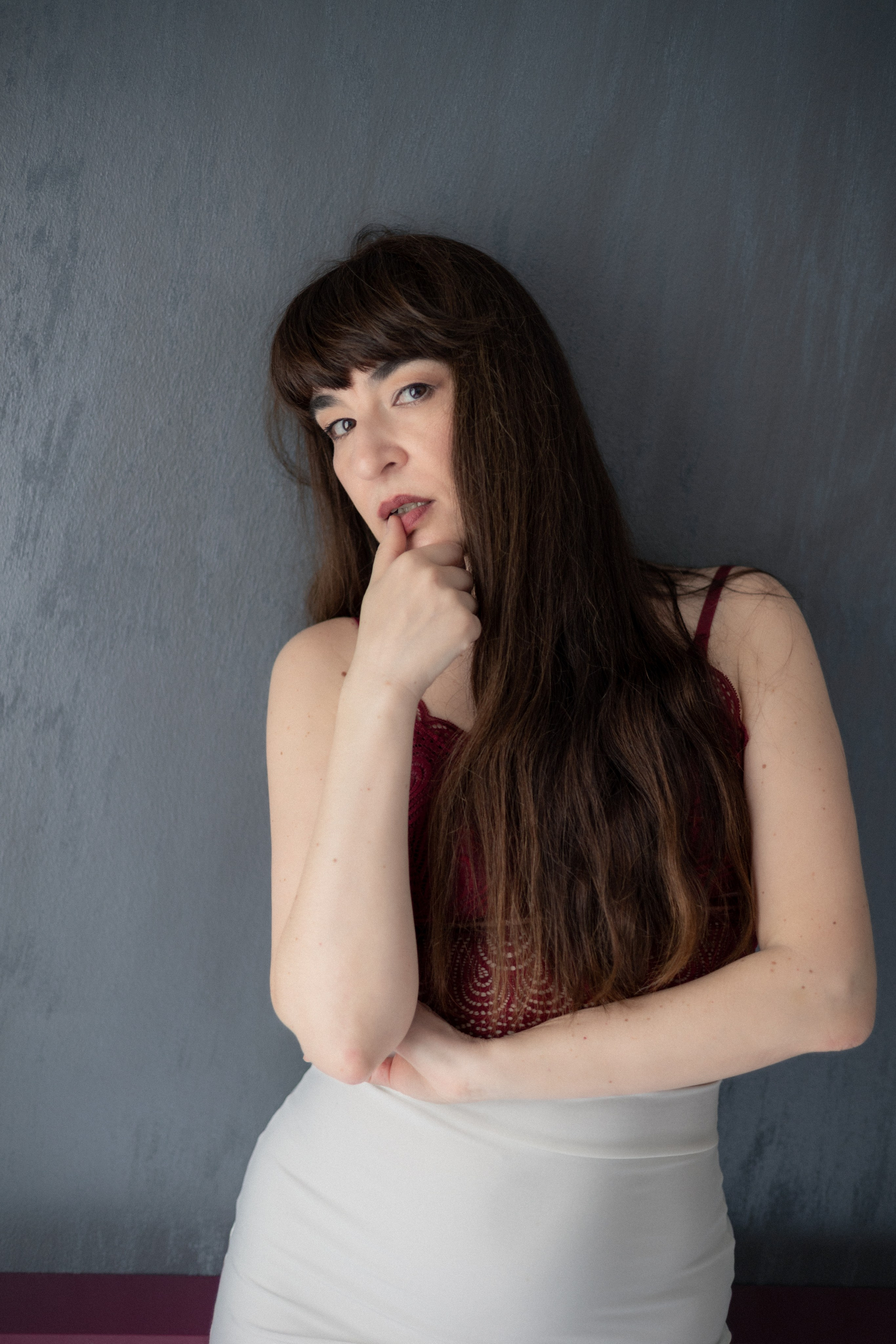 Женщина в кружевном топе стоит у серой стены и касается губ рукой, выразительный портрет с мягким естественным светом