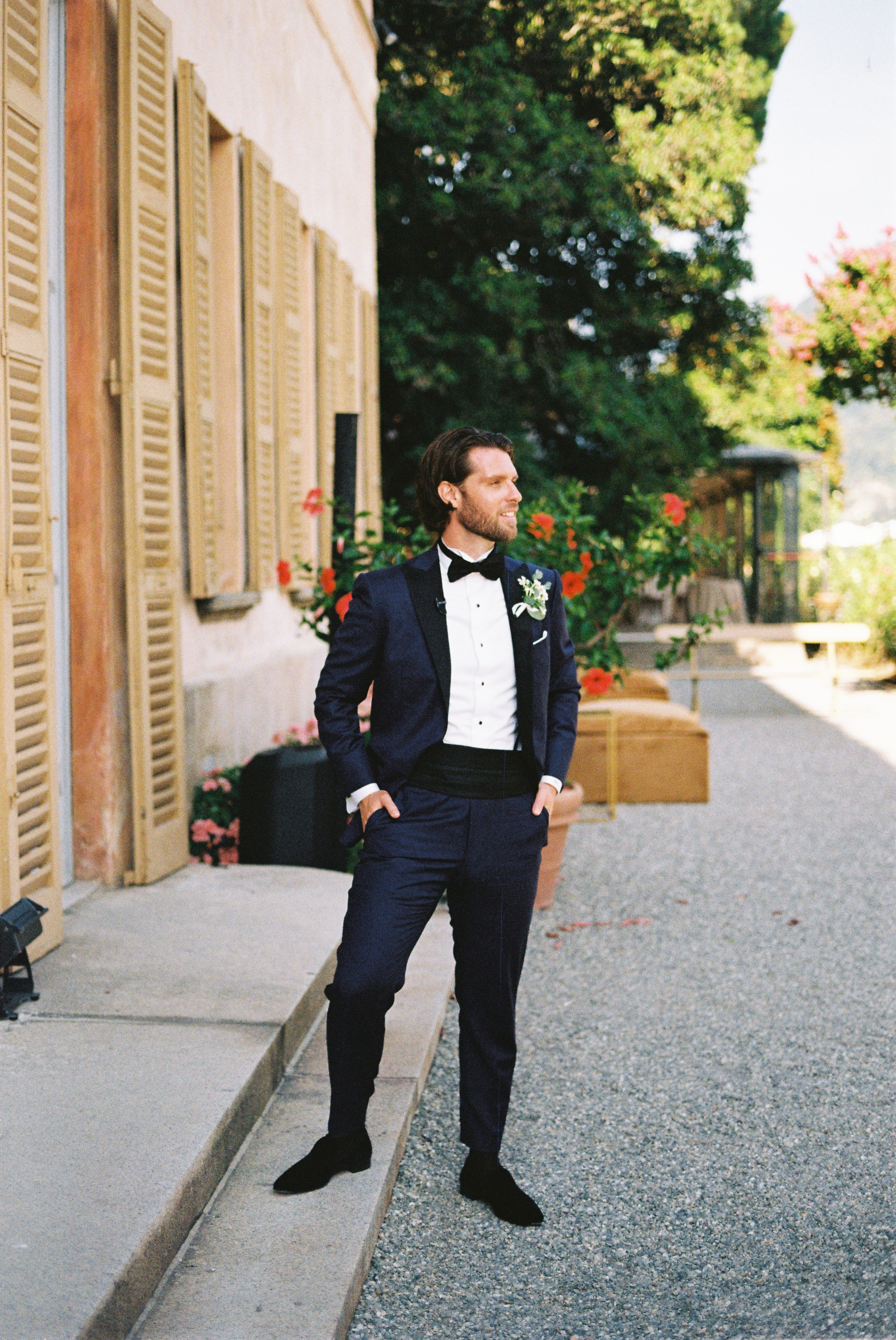Groom in dark suit poses confidently outdoors beside a rustic wall with wooden shutters.