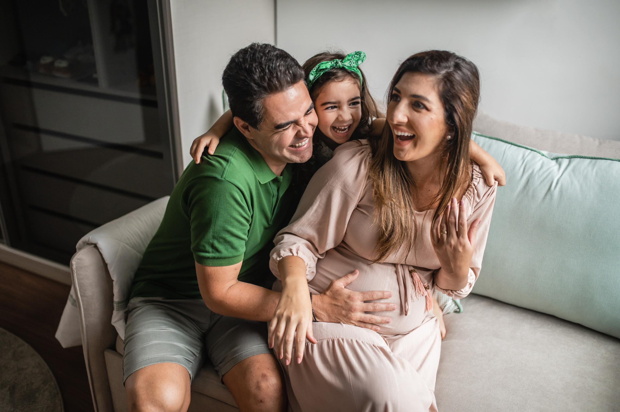Ensaio gestante em família em Juiz de Fora, pai, mãe grávida e filha rindo juntos no sofá