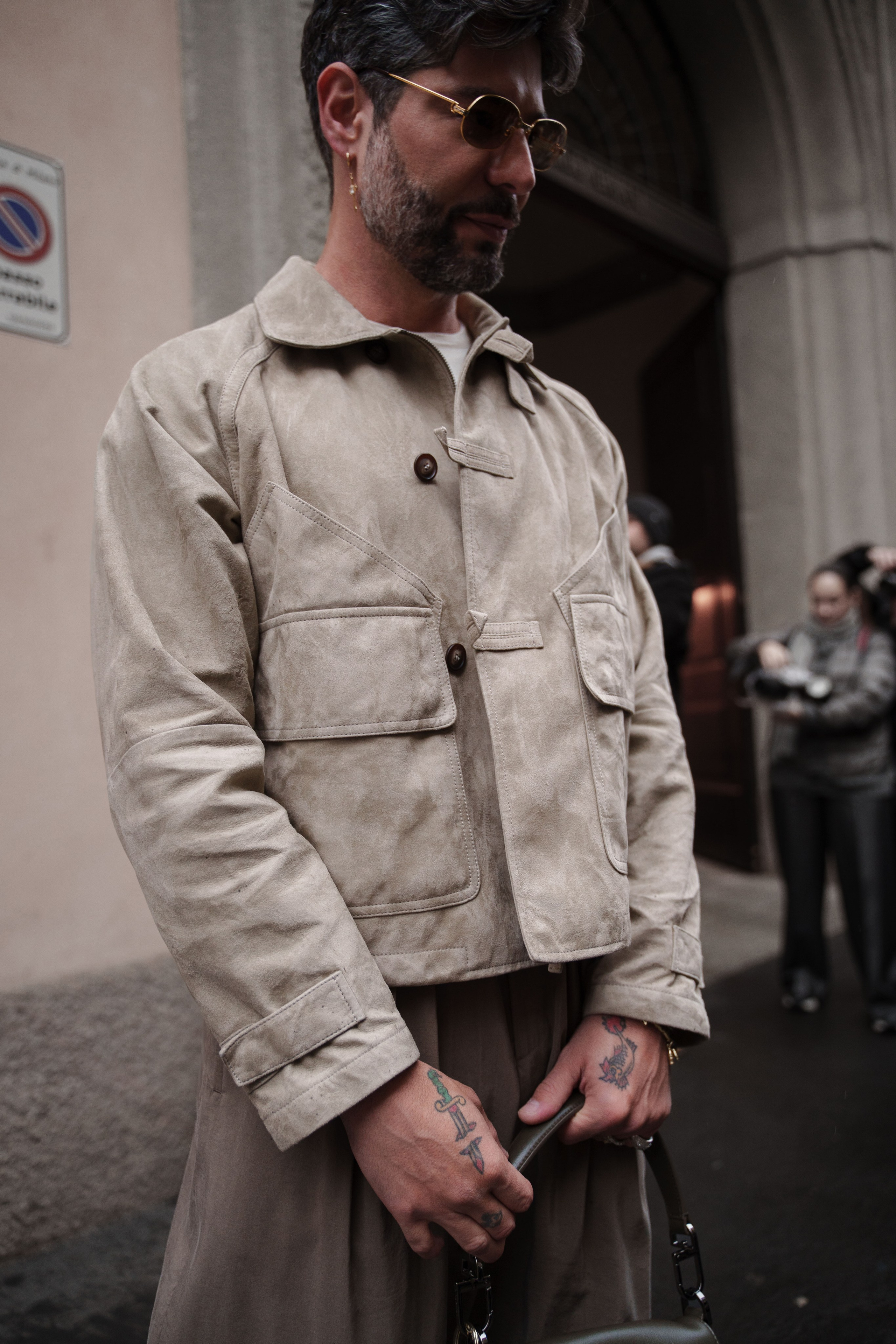 Milan Fashion Week photographer captures a man in a beige suede jacket with tattoos and sunglasses on the streets of Milan