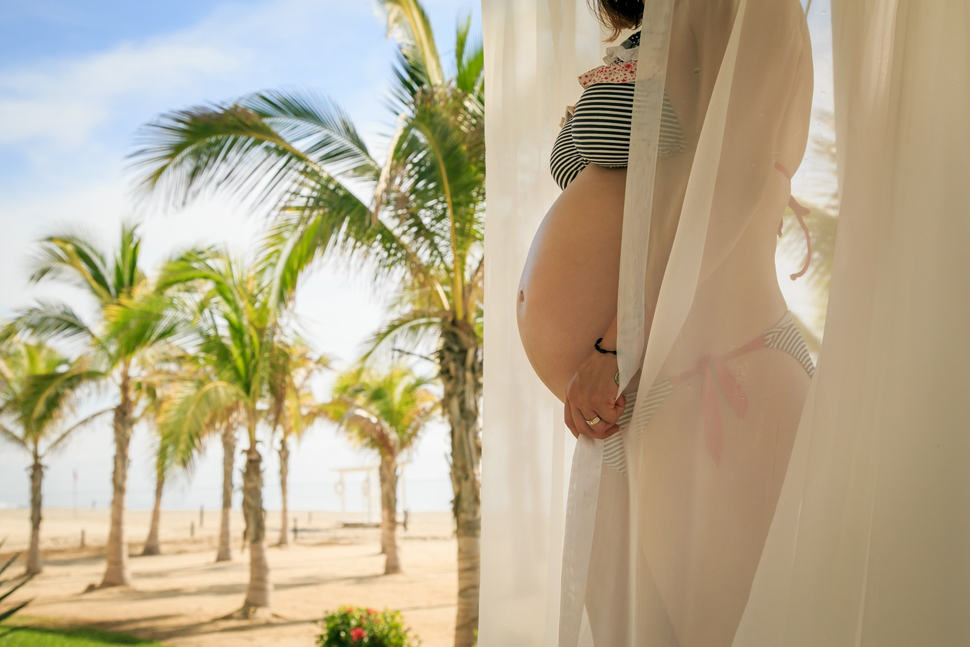 Maternity photoshoot in Los Cabos – close-up of baby bump draped in soft fabric inside a beachfront gazebo, with palm trees and ocean in the background, captured by maternity photographer in Baja California Sur.