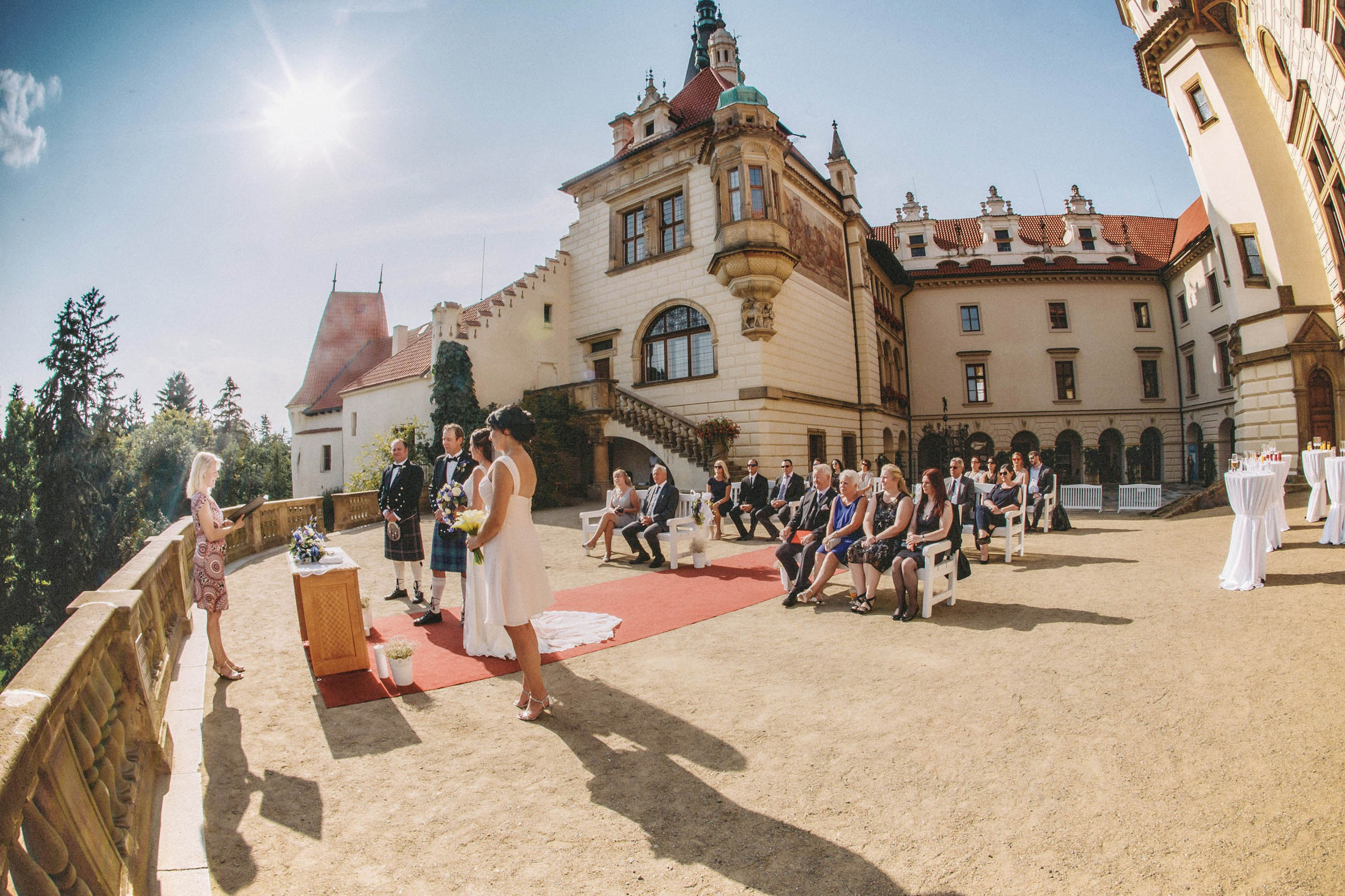 A wide-angle lens captures the atmosphere of an outdoor wedding under a cloudless sky at the historic Pruhonice Castle.