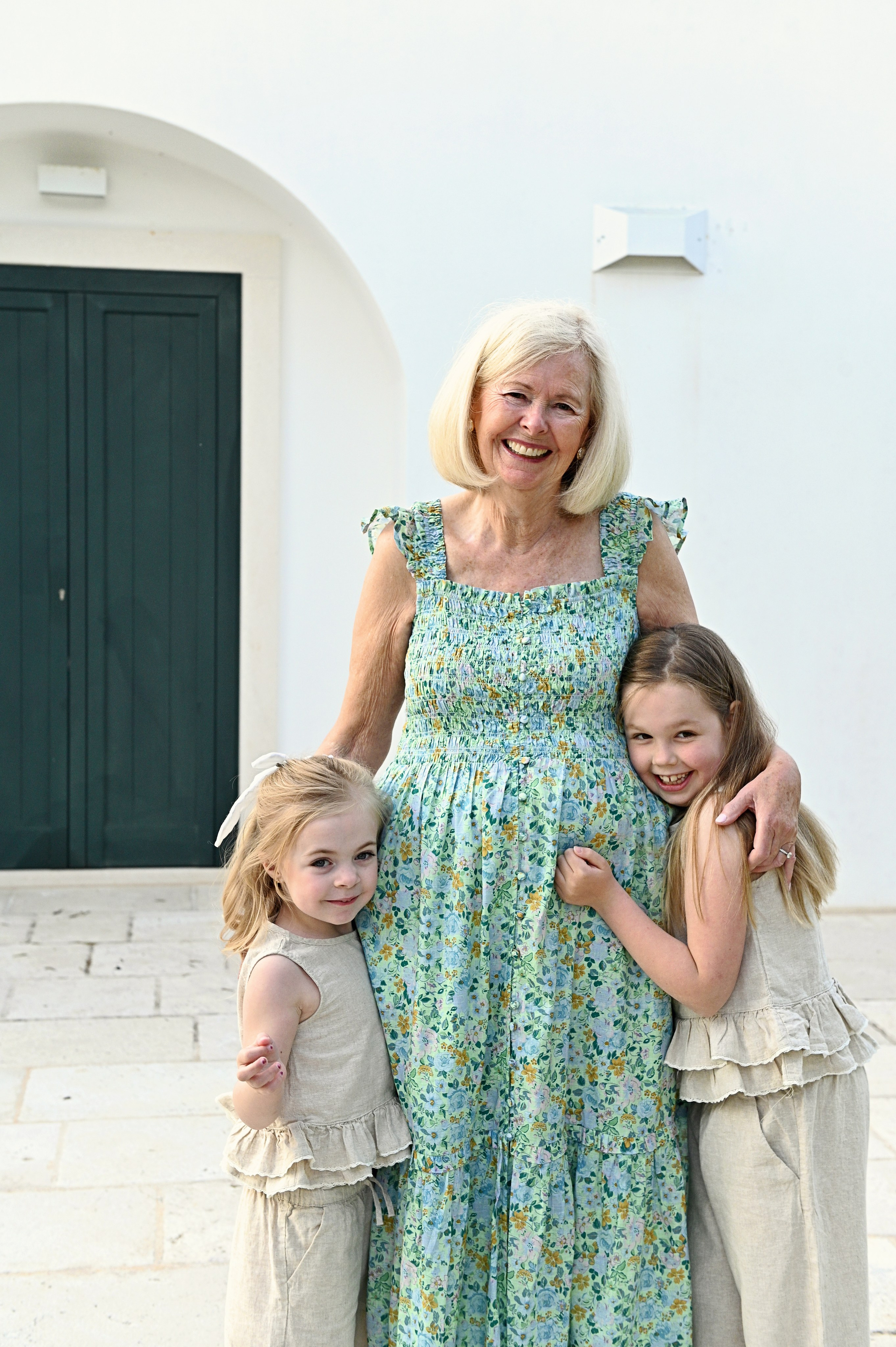 Servizio fotografico di famiglia in Puglia: una nonna sorridente in un abito azzurro a fiori abbraccia le sue due nipotine in completi di lino crema davanti alla parete bianca di un trullo.