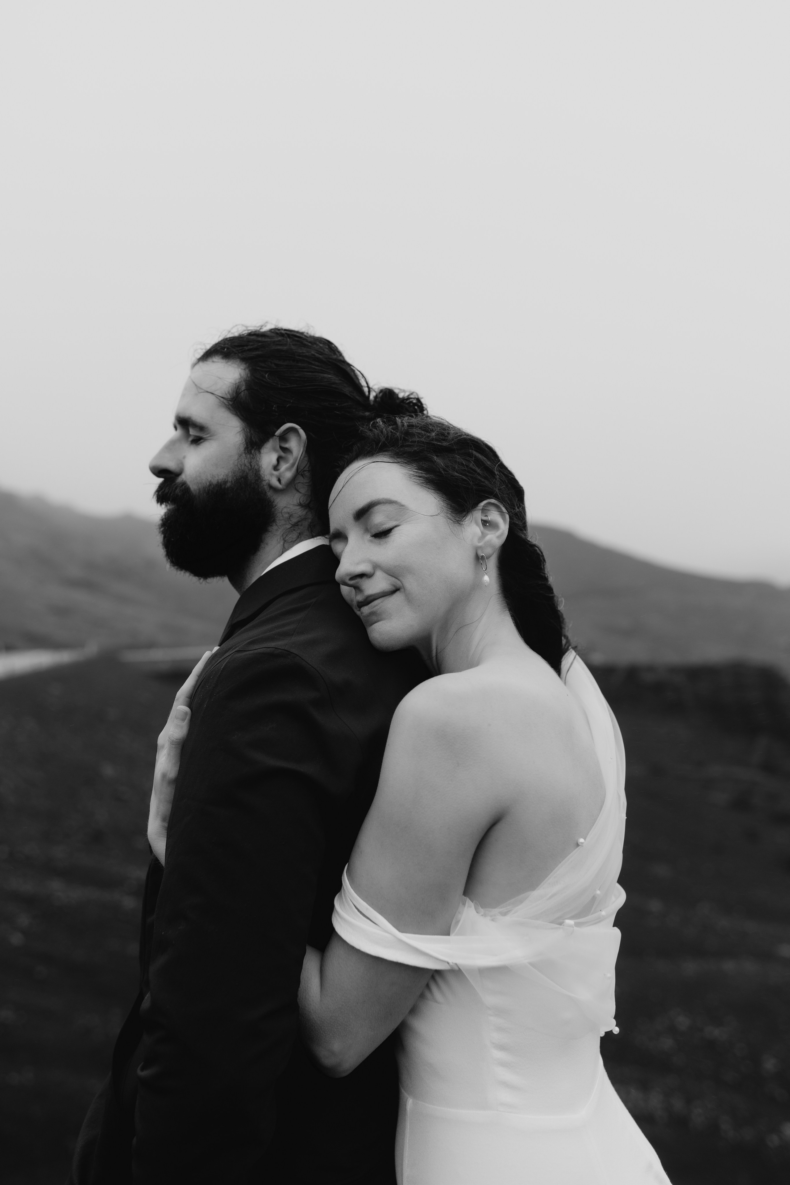 Bride and groom looking out over the vast, mysterious waters of Kleifarvatn, their wedding outfits contrasting against the dark landscape.