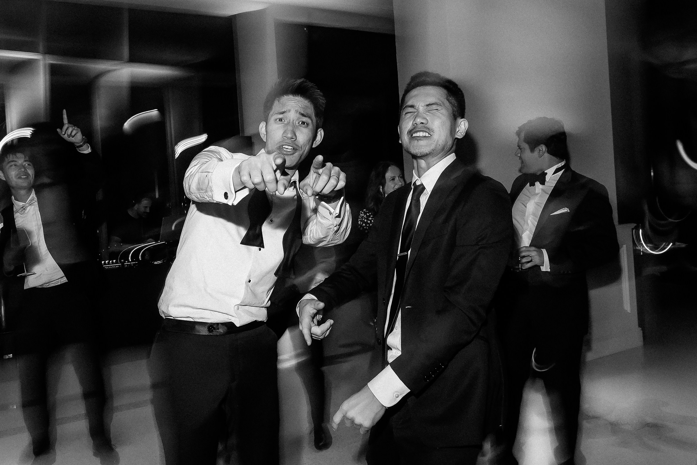 Two men in suits pointing at the camera while dancing, surrounded by friends and a celebratory atmosphere.