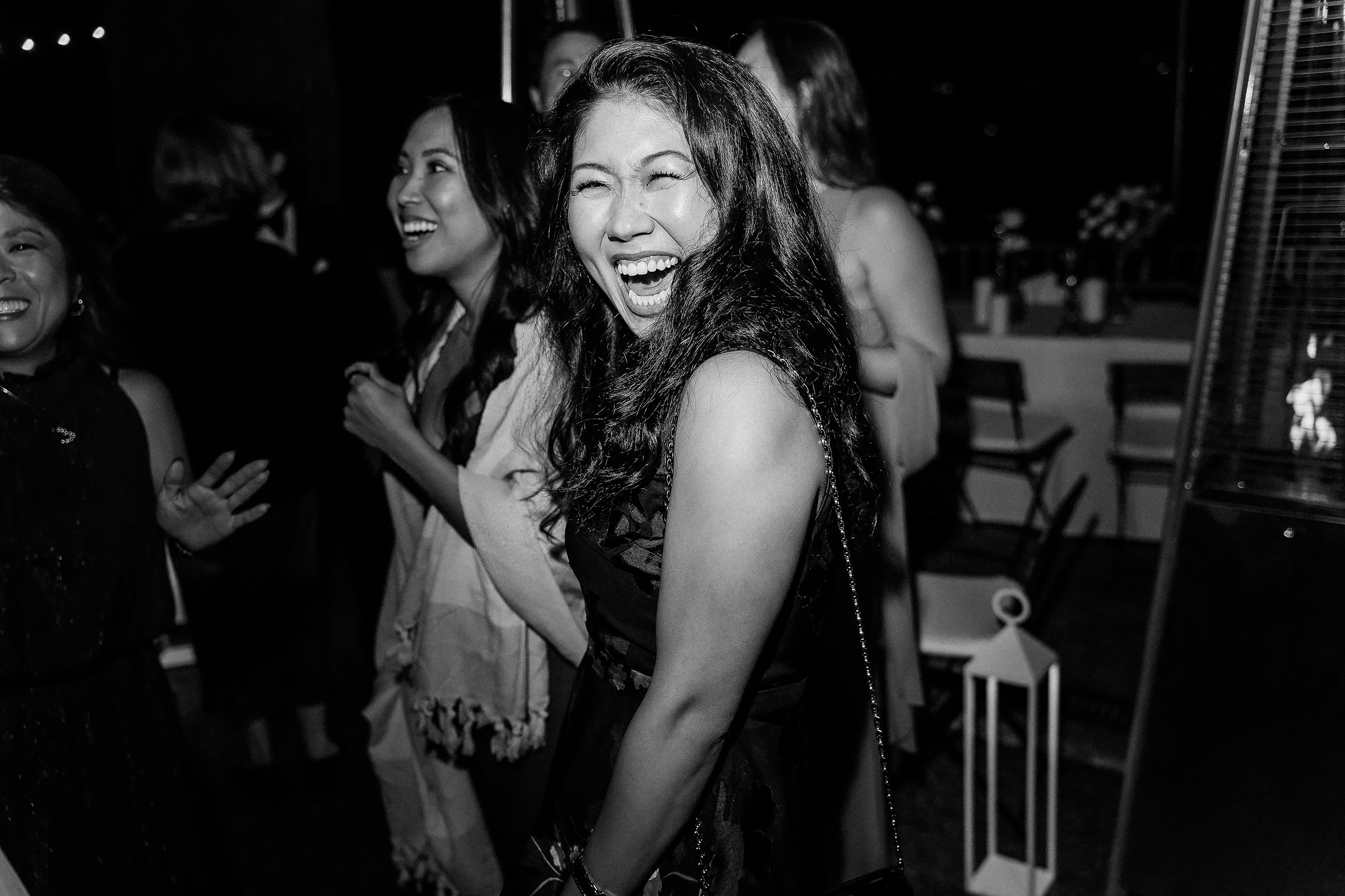 A woman laughing joyfully, surrounded by friends at an evening gathering, with soft lighting enhancing the lively atmosphere.