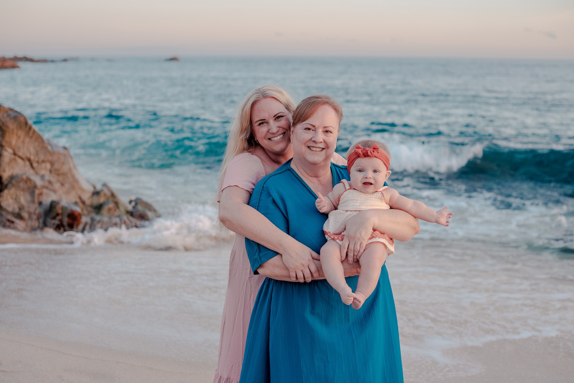 Grandmother mother and baby girl embracing and posing together during family vacation photo session at Playa Monumentos Cabo San Lucas with El Arco in the background