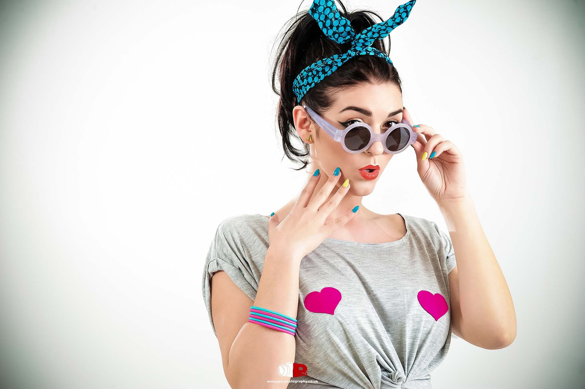 A stylish, retro-fashion woman in a heart-print T-shirt, round sunglasses, and a blue polka dot headband strikes a surprised pose.