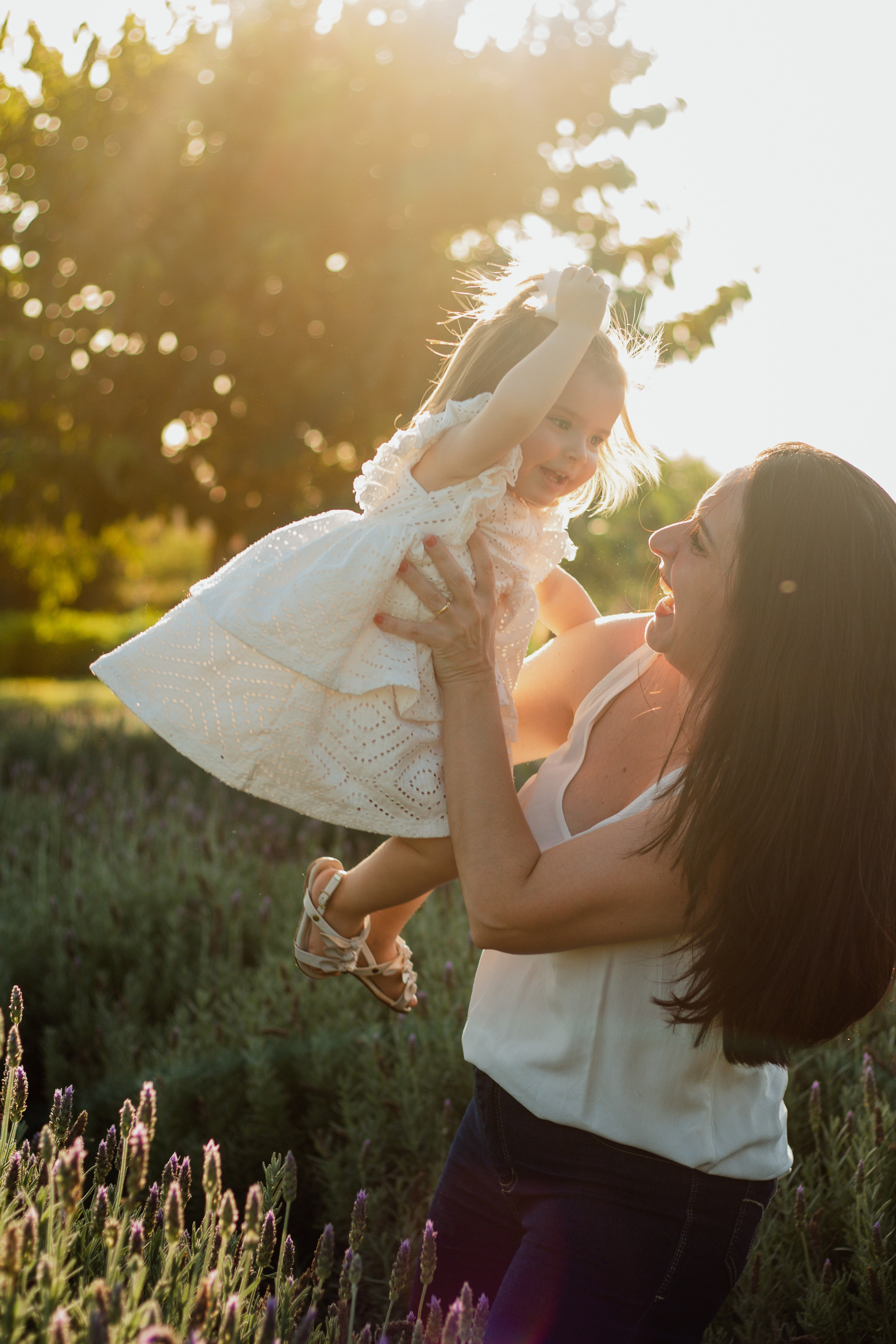 Fotografia espontânea de mãe com blusa branca e calça jeans, e filha com vestido branco e laço no cabelo, em campo de lavanda ao pôr do sol, em Holambra.