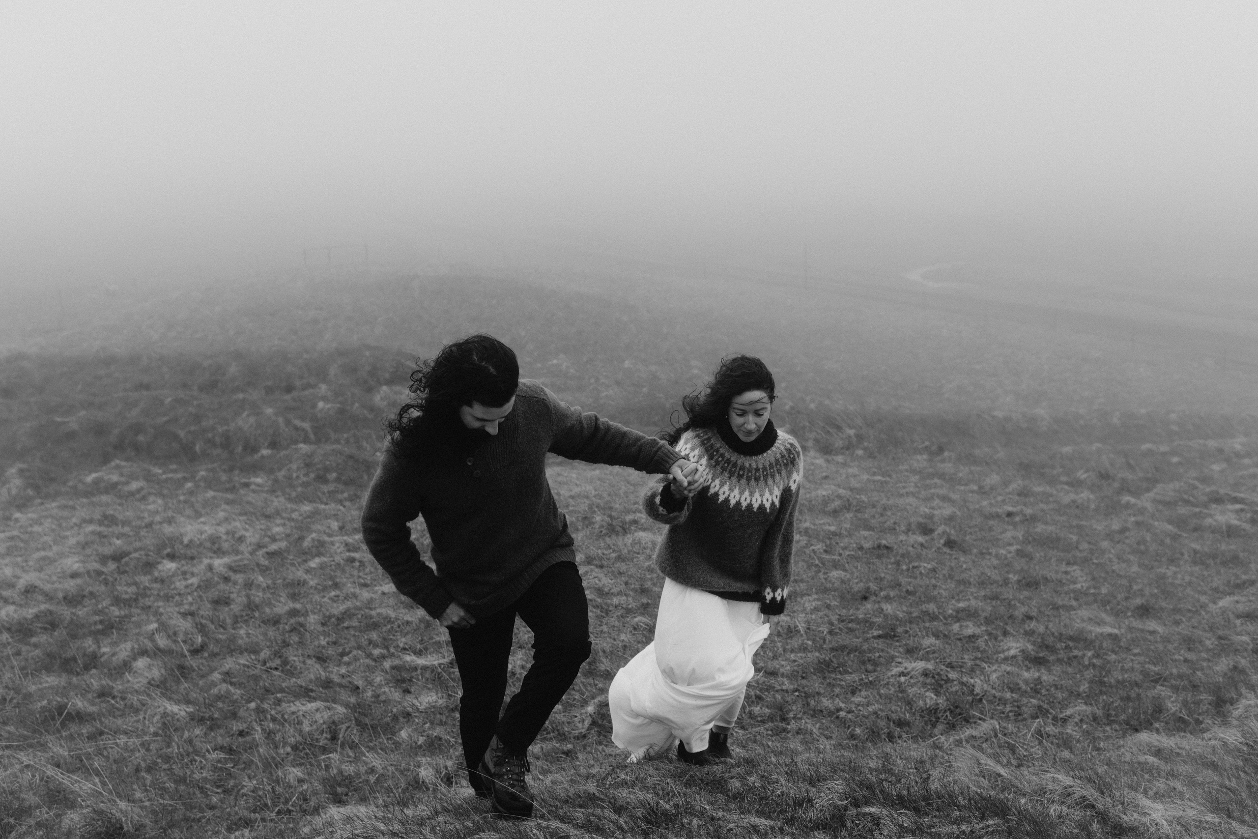 Bride and groom sitting on the stone wall near Krýsuvíkurkirkja, wrapped in a cozy Icelandic blanket, as the stormy sky looms above.