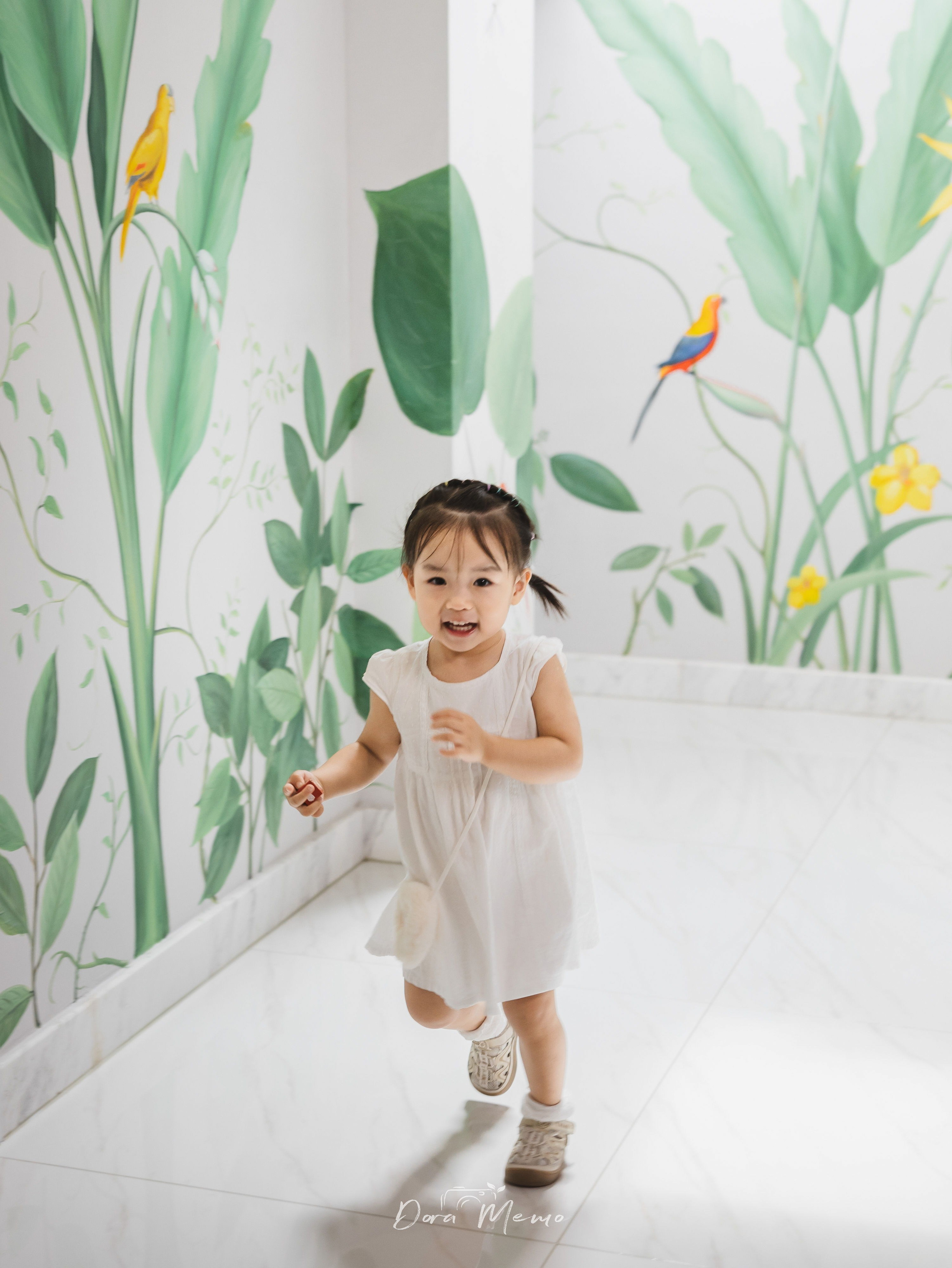 A little girl in a white dress smiles in front of a painted wall inside her family home.