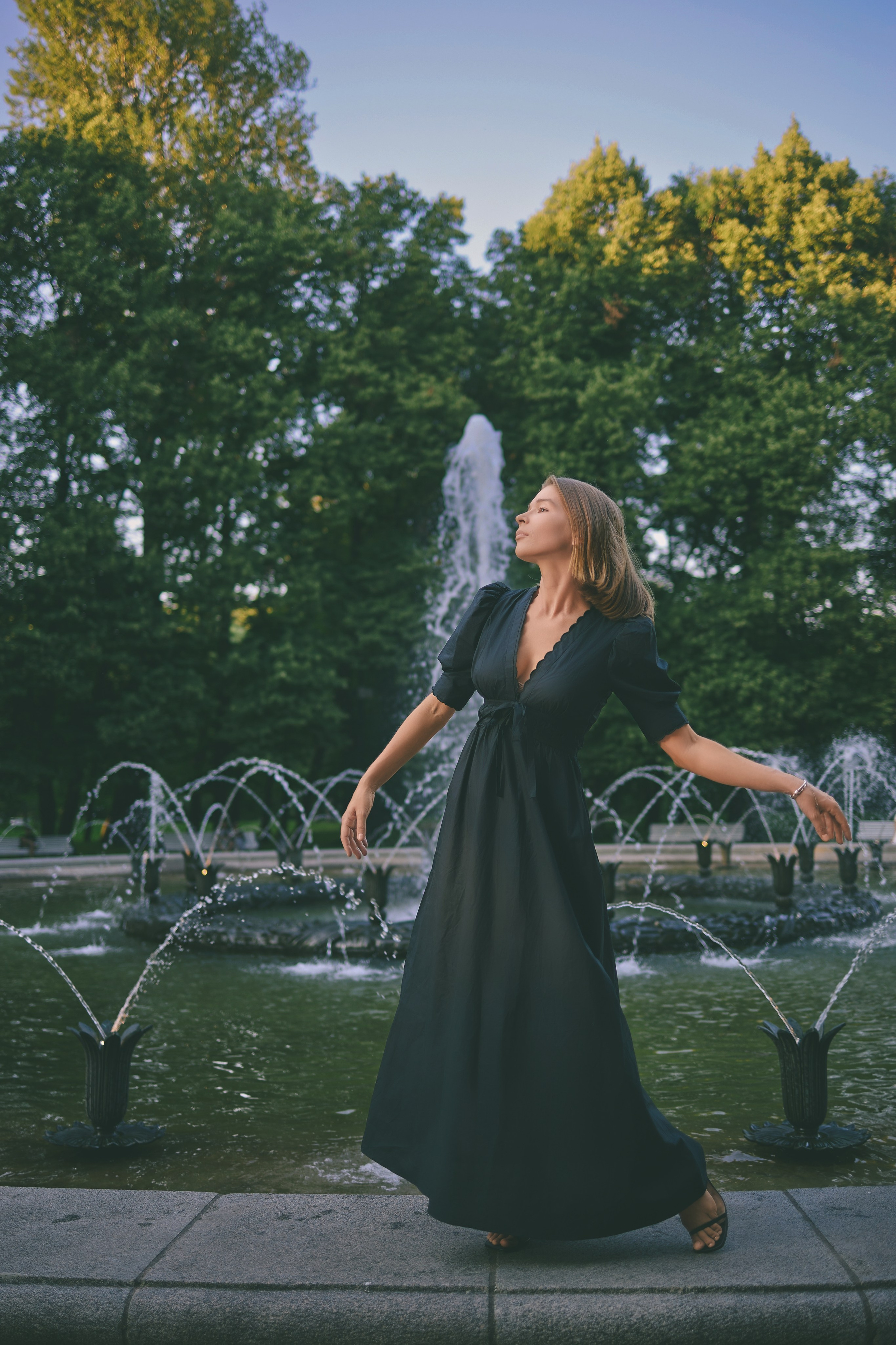 Уличная фотосесия девушка в черном платье танцует у фонтана