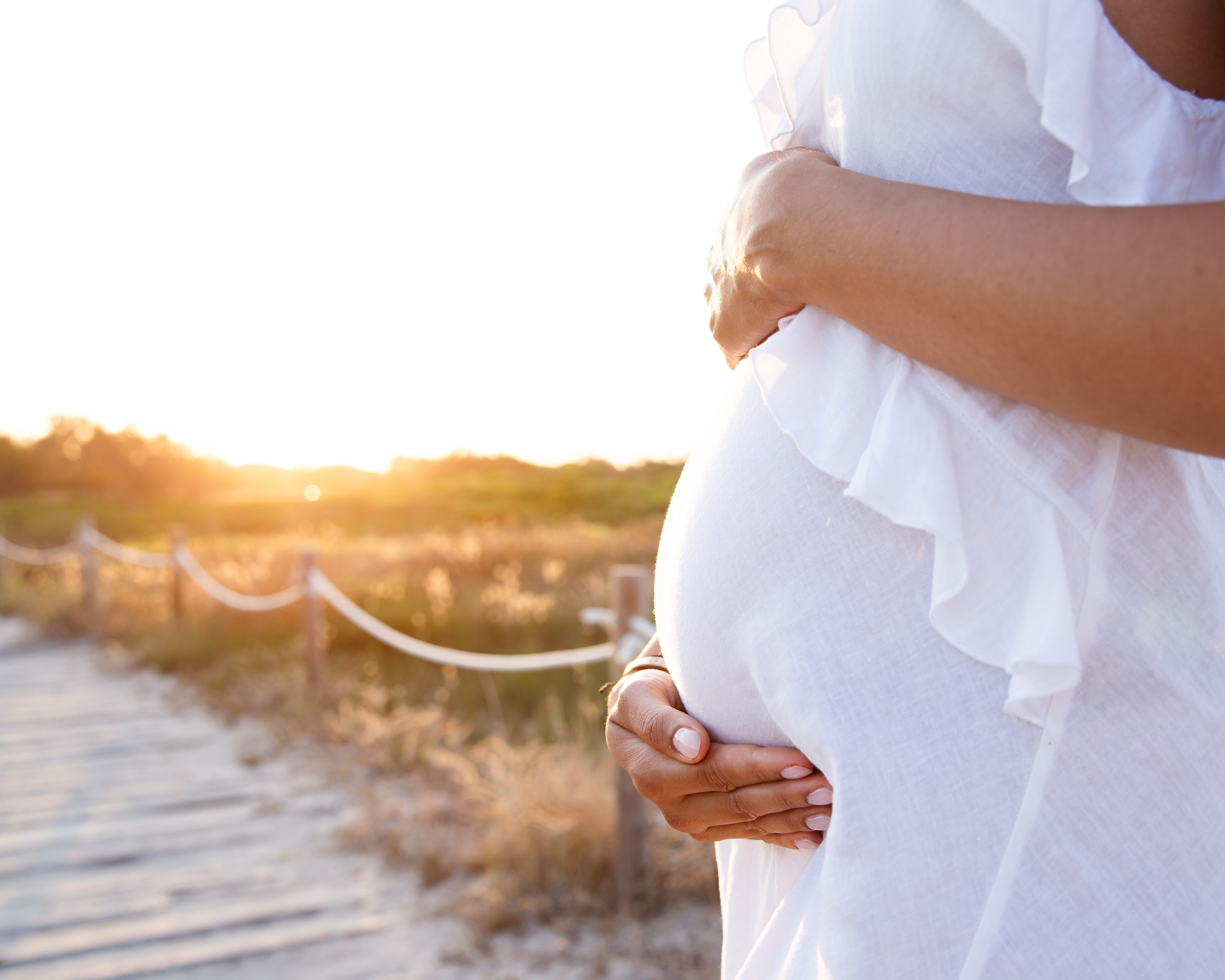 Primer plano de embarazo en Jávea, España — manos tiernas abrazando suavemente una barriga al atardecer sobre una pasarela. Perfecta para quienes buscan fotos íntimas y emocionales de maternidad en Jávea y la Costa Blanca. 