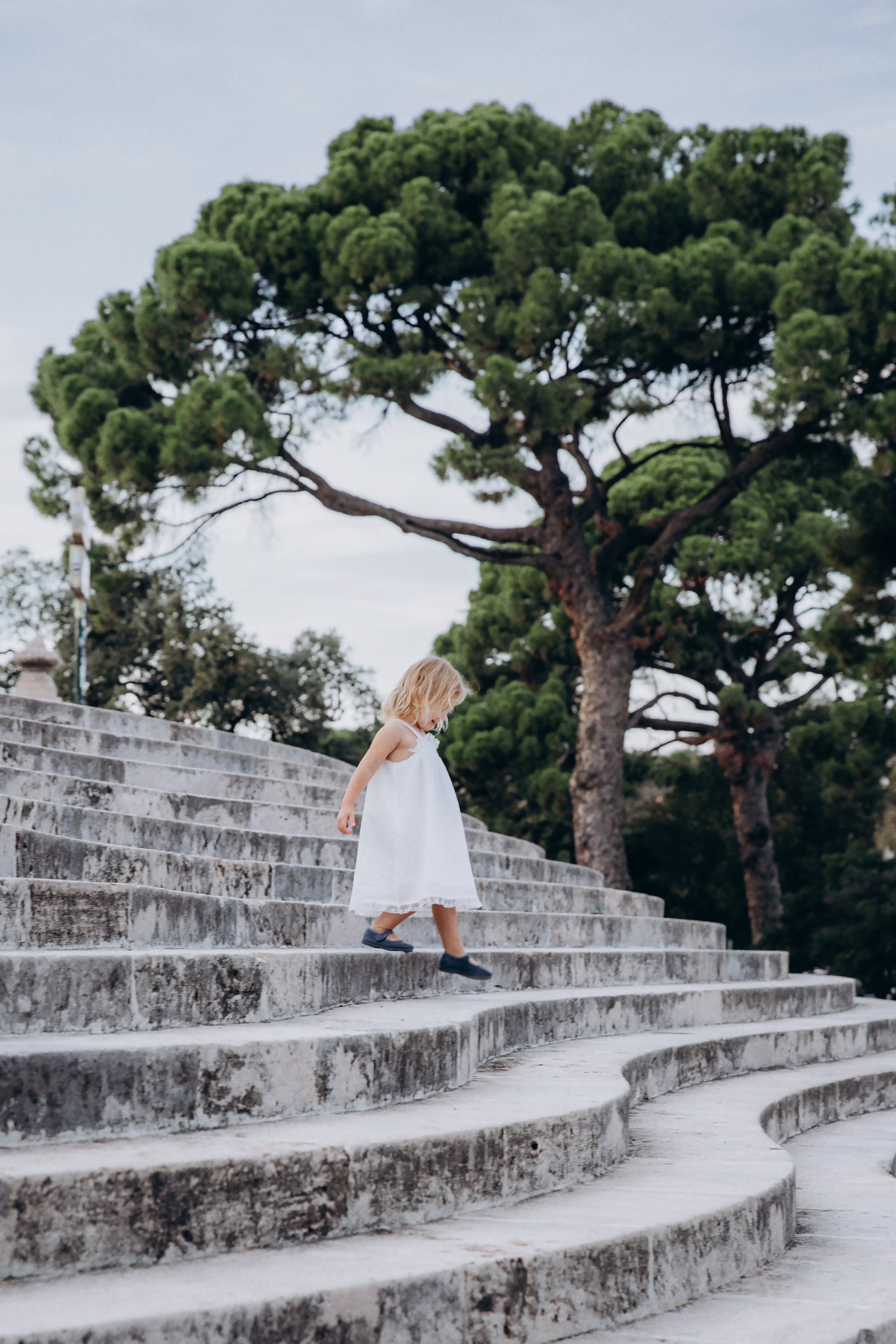 Niña bajando por escalones de piedra bajo frondosos pinos verdes durante una tranquila sesión de fotos familiar en Valencia, España — un momento perfecto para quienes buscan fotografía familiar natural y artística en Valencia y en toda España. Ideal para familias que desean recuerdos sinceros y atemporales en entornos al aire libre.