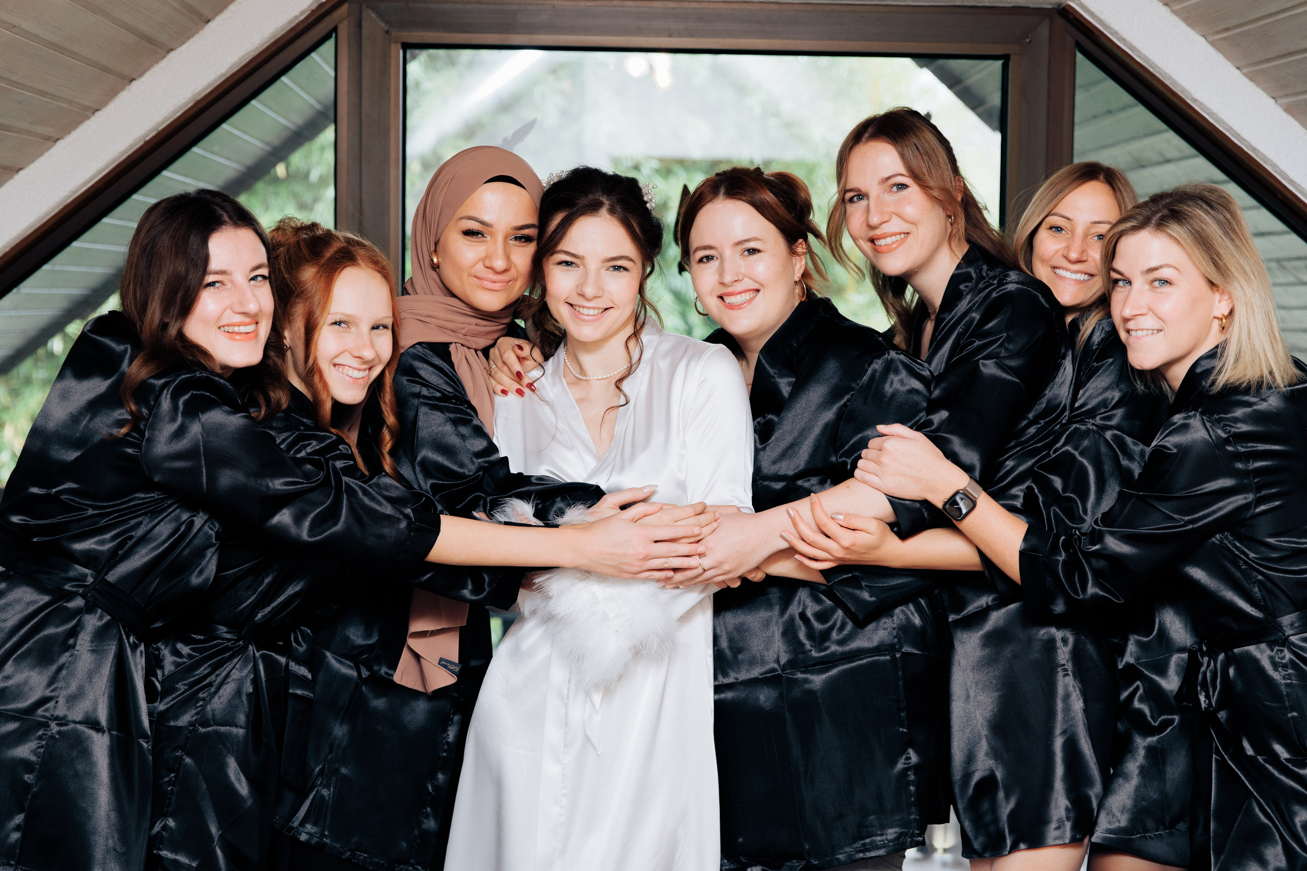Die Brautjungfern in schwarzen Seidenmorgenmänteln umarmen die Braut, die in einem weißen Morgenmantel in der Mitte steht. Alle lachen und schauen in die Kamera