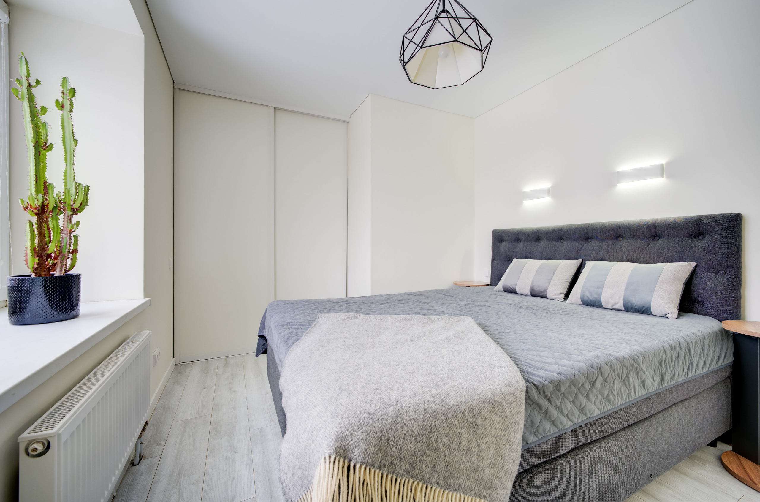 Miegamasis su minkštu galvūgaliu, sieniniais šviestuvais ir dideliu kaktusu prie lango.