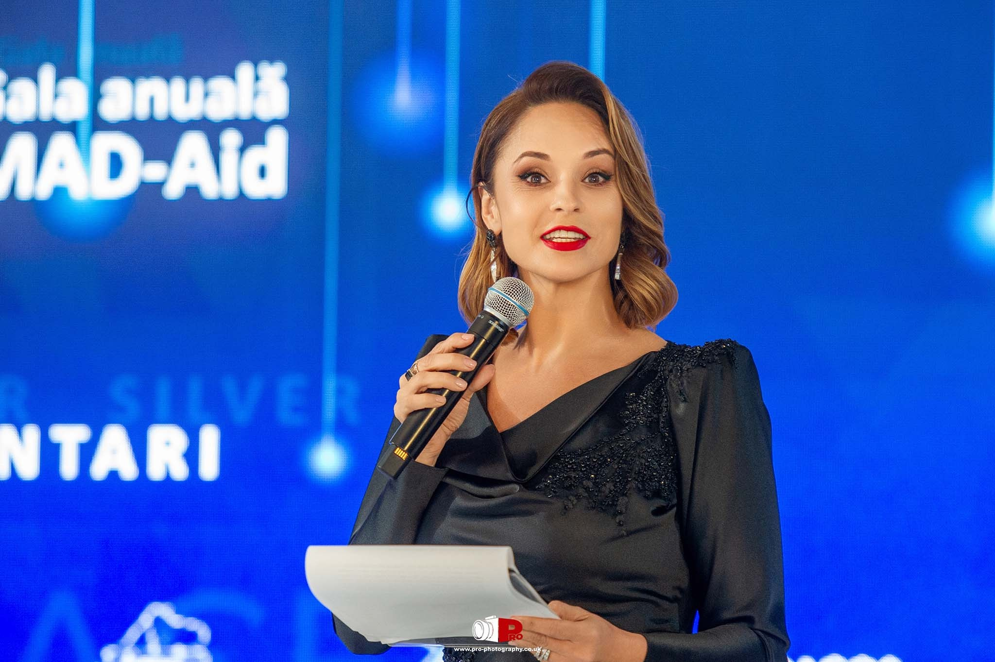 A female speaker passionately addresses the audience at the MAD-Aid Gala event, holding a microphone with a captivating background.