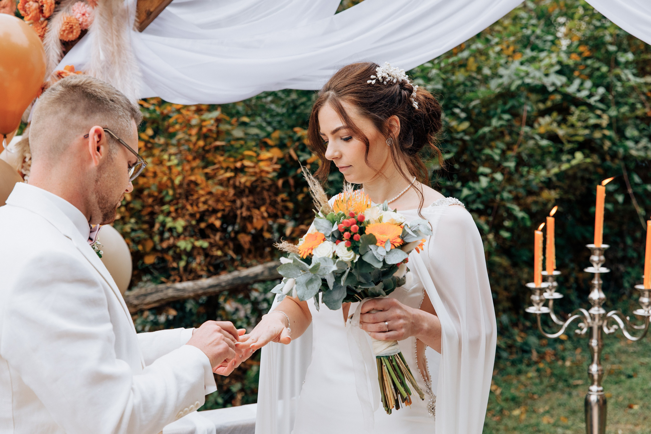 Der Bräutigam steckt der Braut den Ehering an, sie hält in ihrer anderen Hand einen schönen Hochzeitsstrauß.