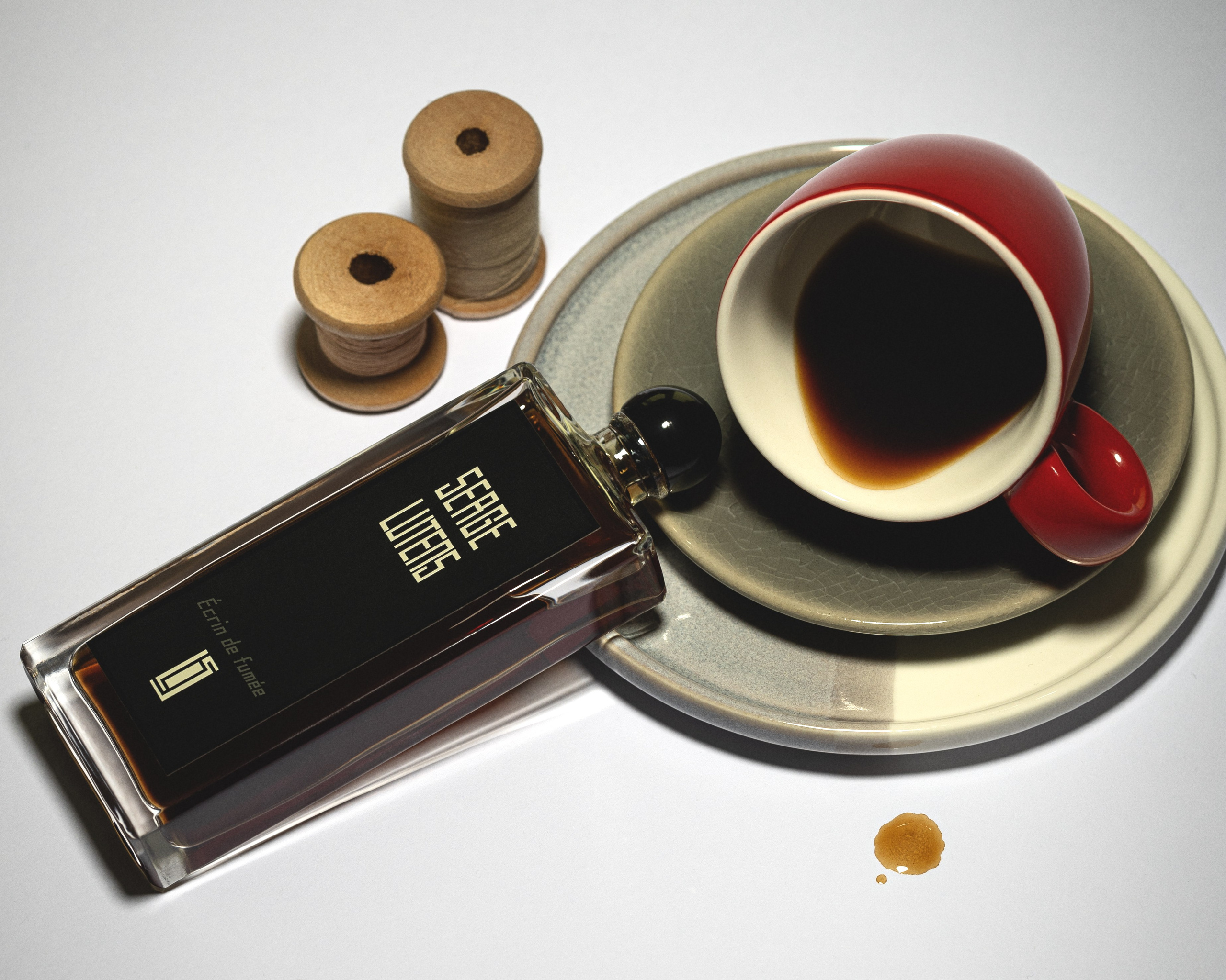 Serge Lutens perfume bottle with a black label placed next to a red coffee cup on a ceramic plate, creating a stylish composition with coffee drops and wooden spools in the background. Luxury fragrance photography with warm tones and vintage aesthetics