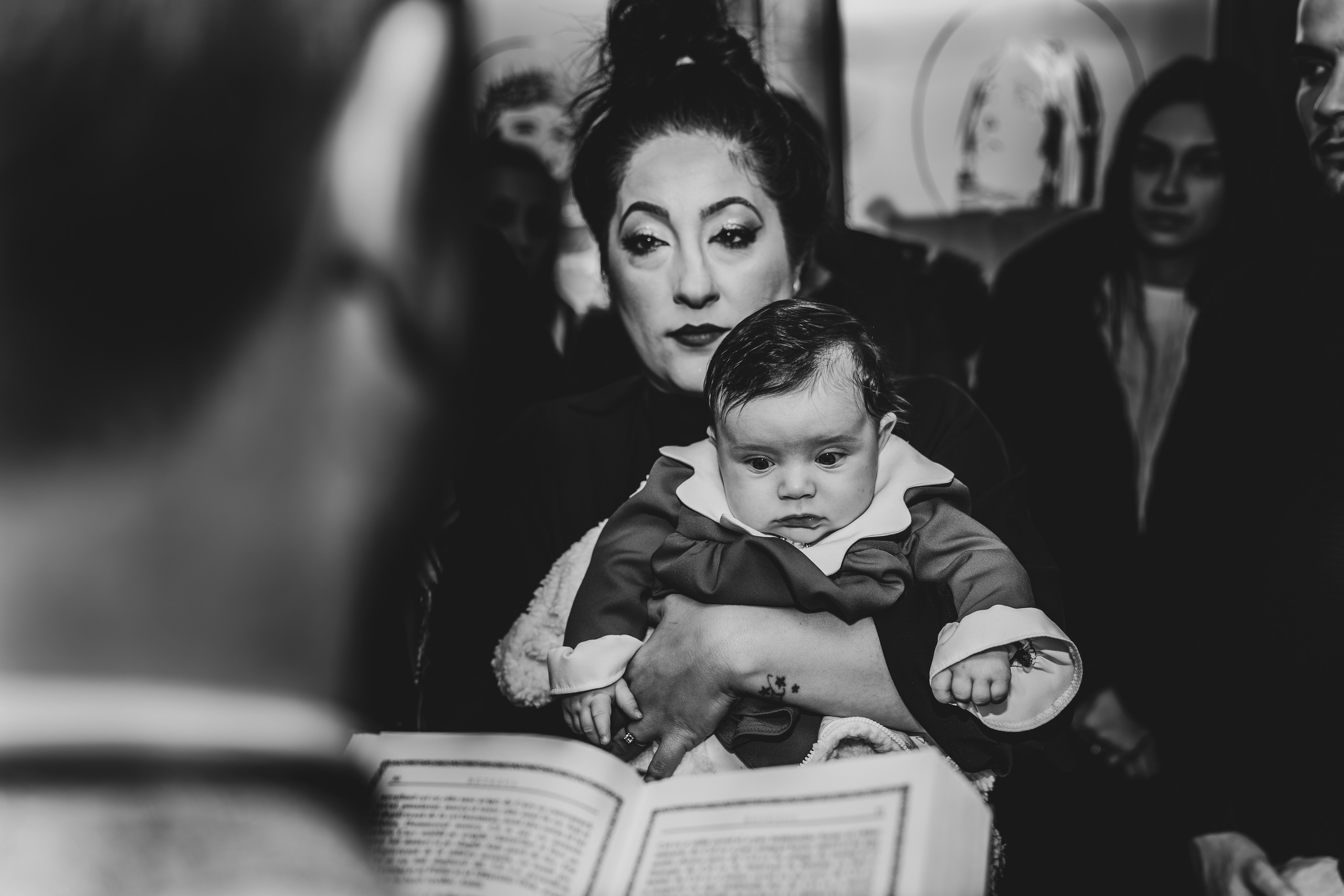 Detaliu intim de la botez: peste umărul preotului, privirea plină de grijă și iubire a nașei către pruncul Eveline Maria, pe care îl ține în brațe în timpul ceremoniei. Fotografie naturală și emoționantă.