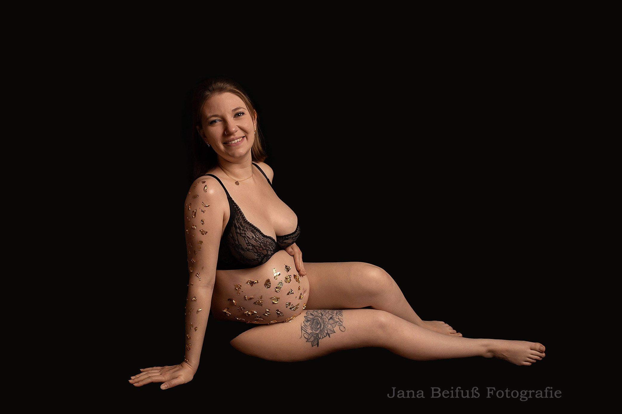 werdende Mama sitzt in schwarzer Unterwäsche mit Goldflocken auf der Haut beim Babybauchshooting bei Jana Beifuß in Weißenhorn bei Ulm - Schwangerschaftsfotografie Weißenhorn bei Ulm