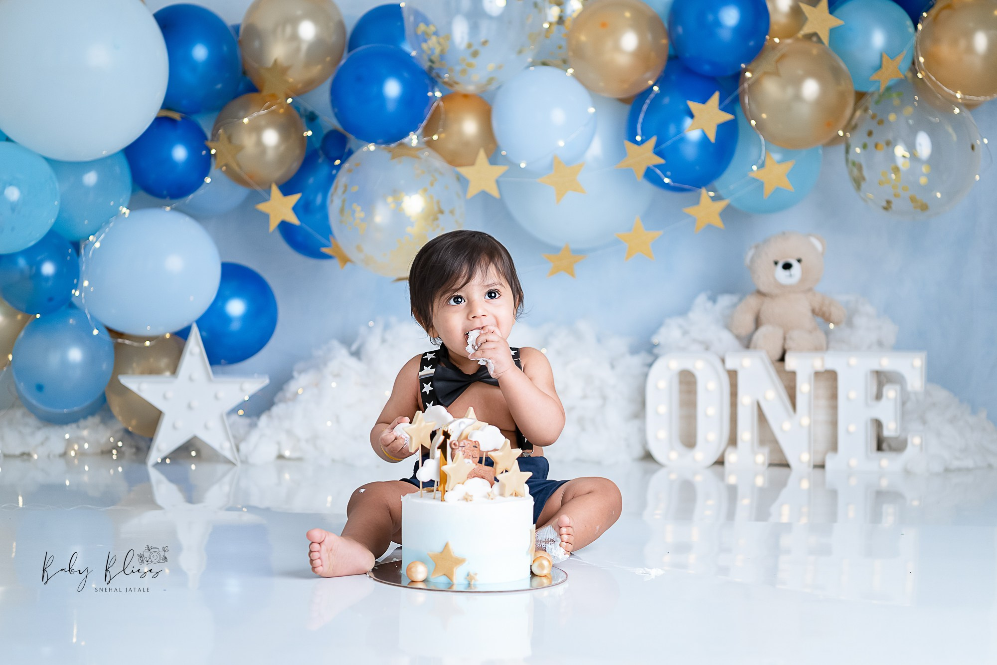 child having cake with blue, gold, light blue colour balloon arch as a background.
