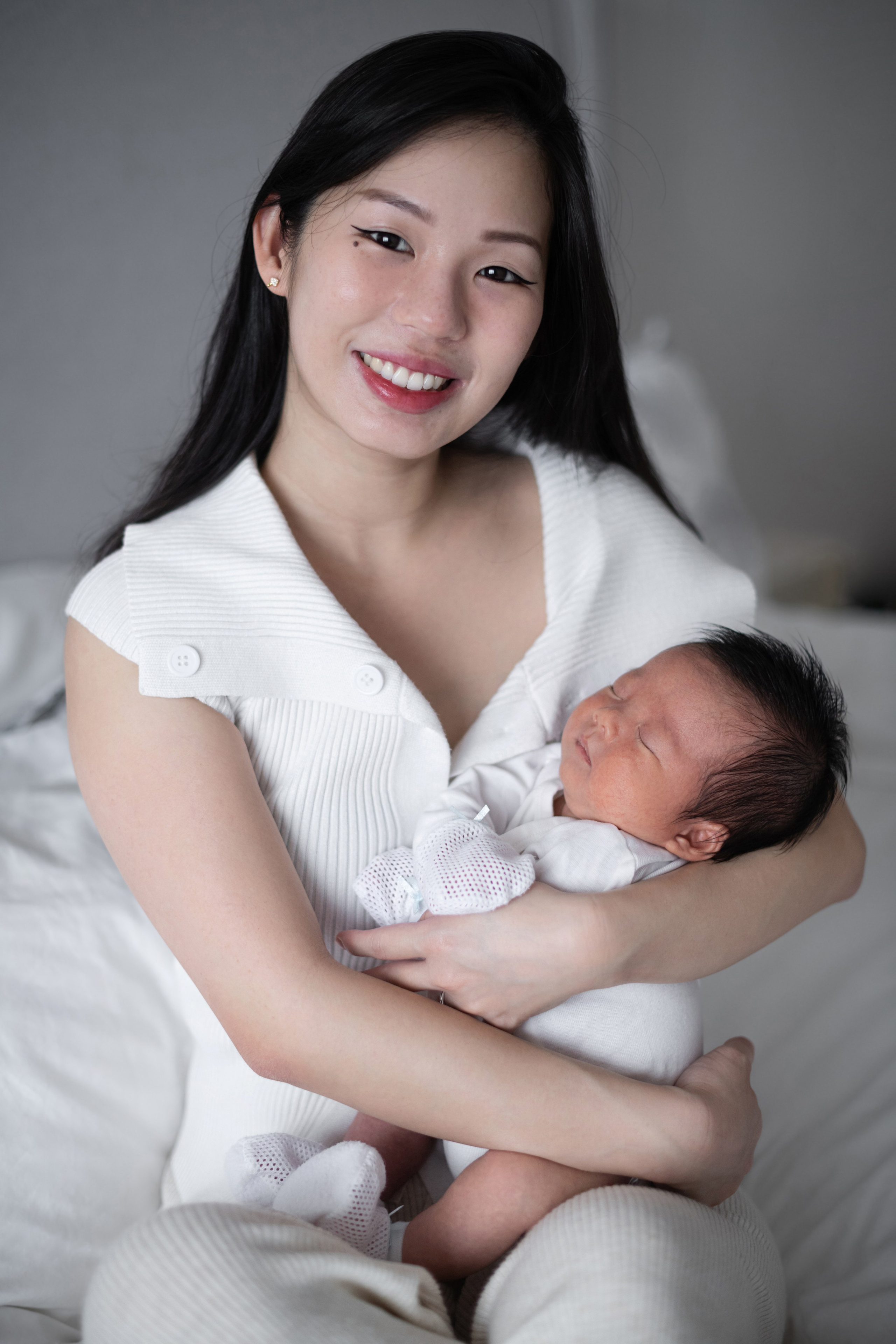 Young Asian mum cradling her swaddled newborn son on the bed, both dressed in white, natural window light.