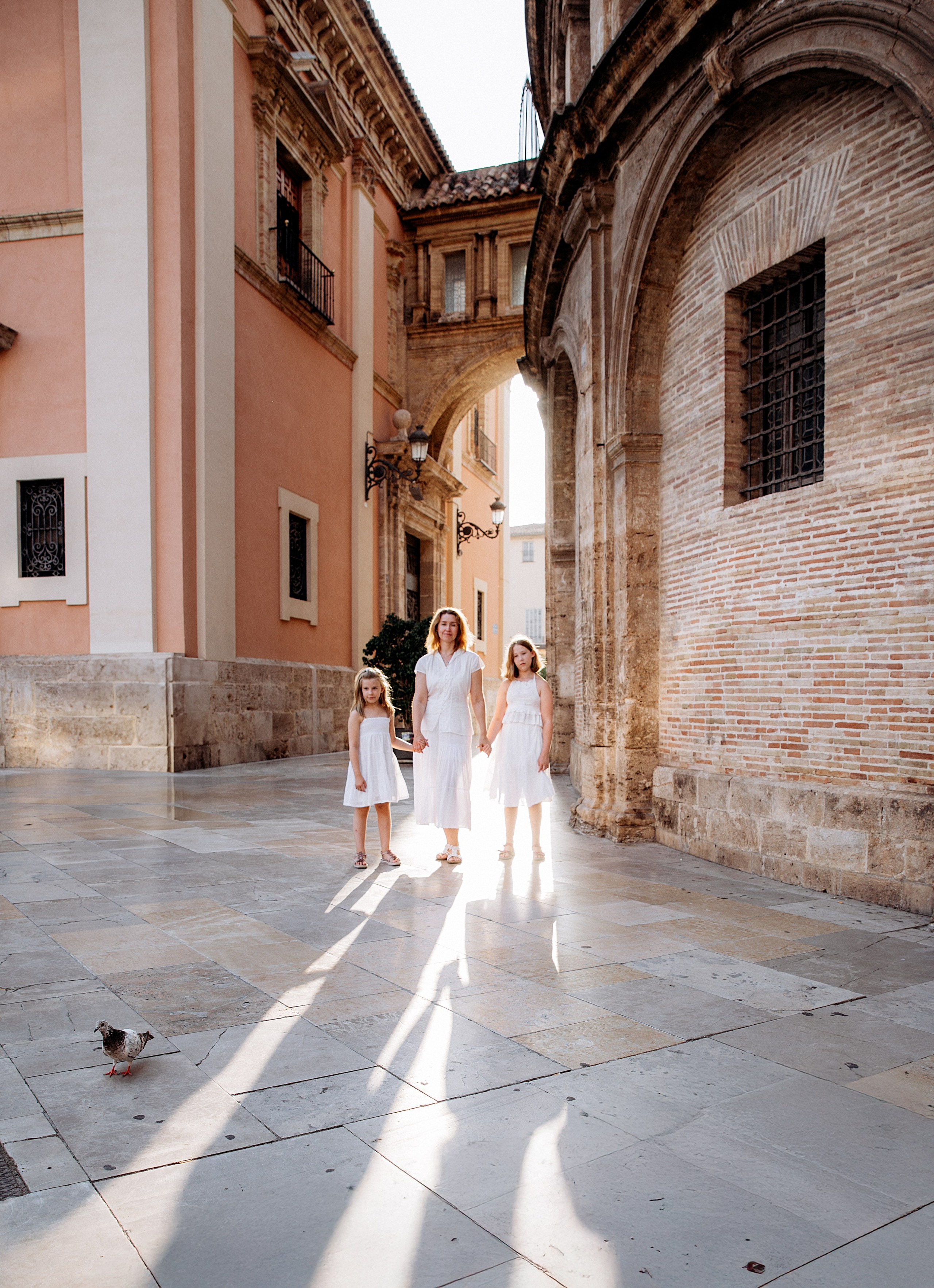 Sesión familiar elegante en el centro histórico de Valencia, España, con una madre y sus dos hijas vestidas de blanco caminando de la mano por calles encantadoras. Inspiración ideal para sesiones familiares atemporales y con estilo en Valencia y toda España.