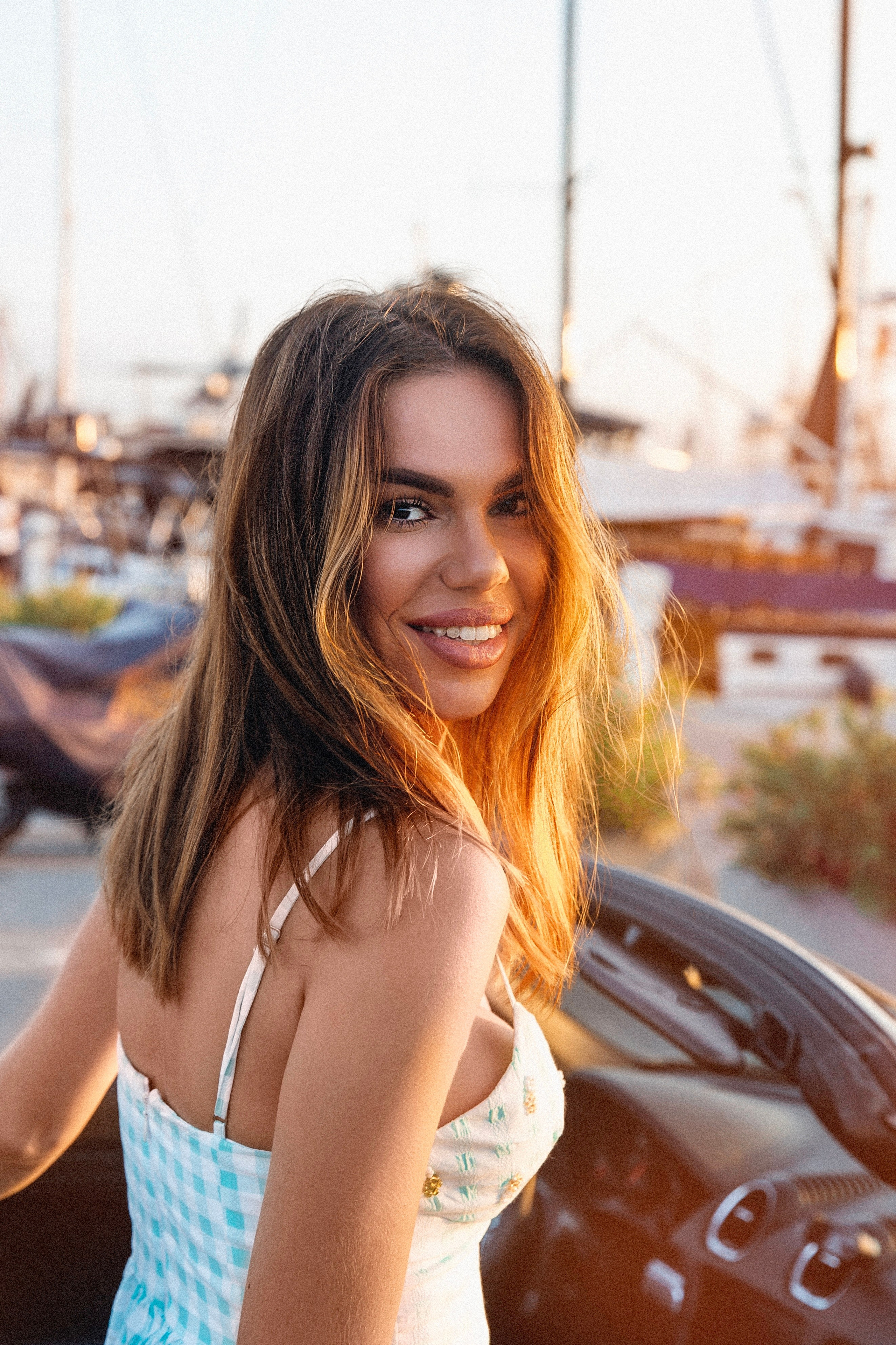 Портрет улыбающейся женщины в голубом и белом летнем платье, стоящей рядом с кабриолетом на закате, с лодками и мачтами на заднем плане на пристани в Валенсии, Испания