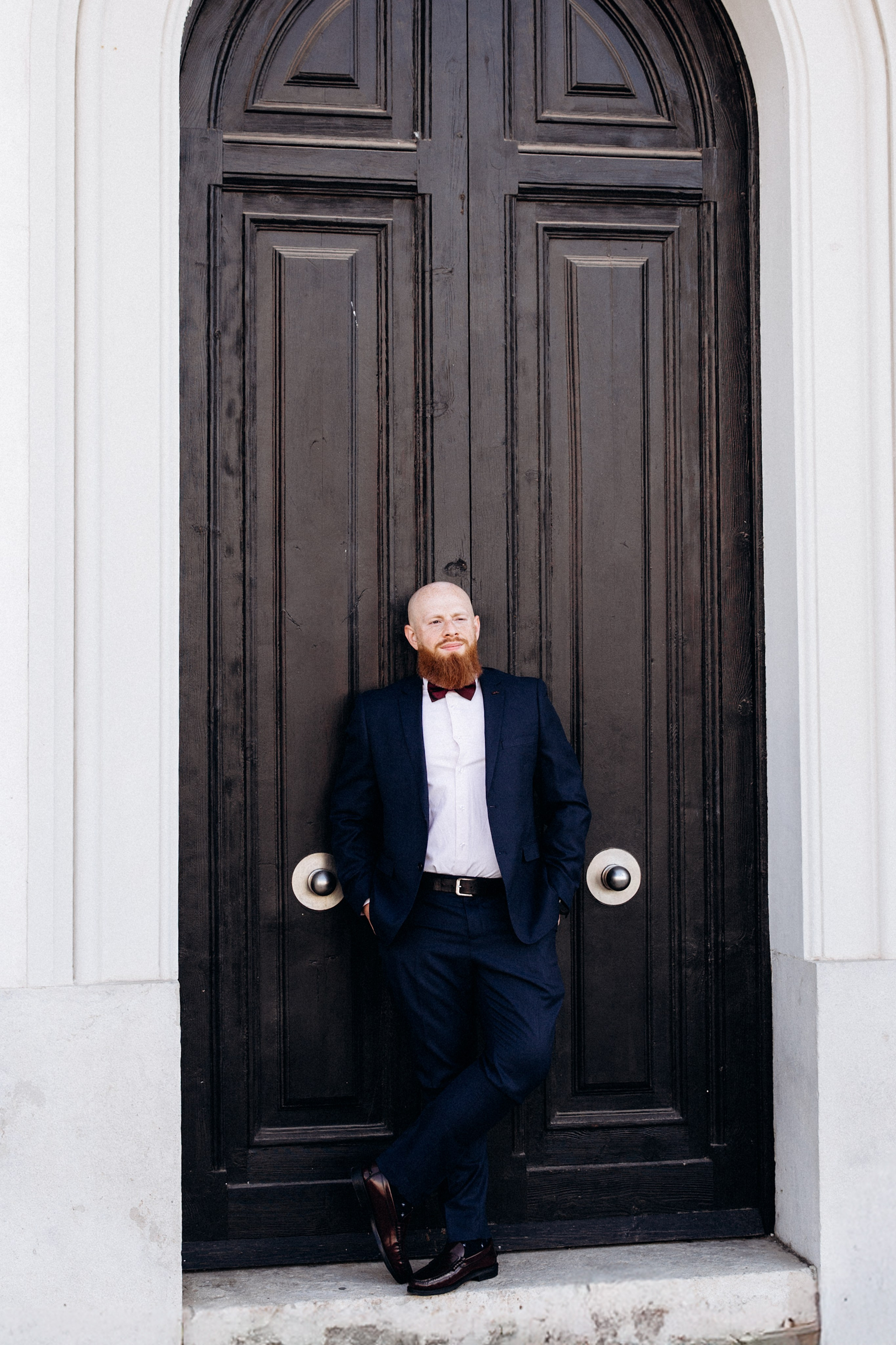 Groom portrait in elegant wedding attire leaning against a grand black door in Madrid, Spain — a stylish and timeless shot perfect for couples seeking artistic and refined wedding photoshoots in Madrid and across Spain.