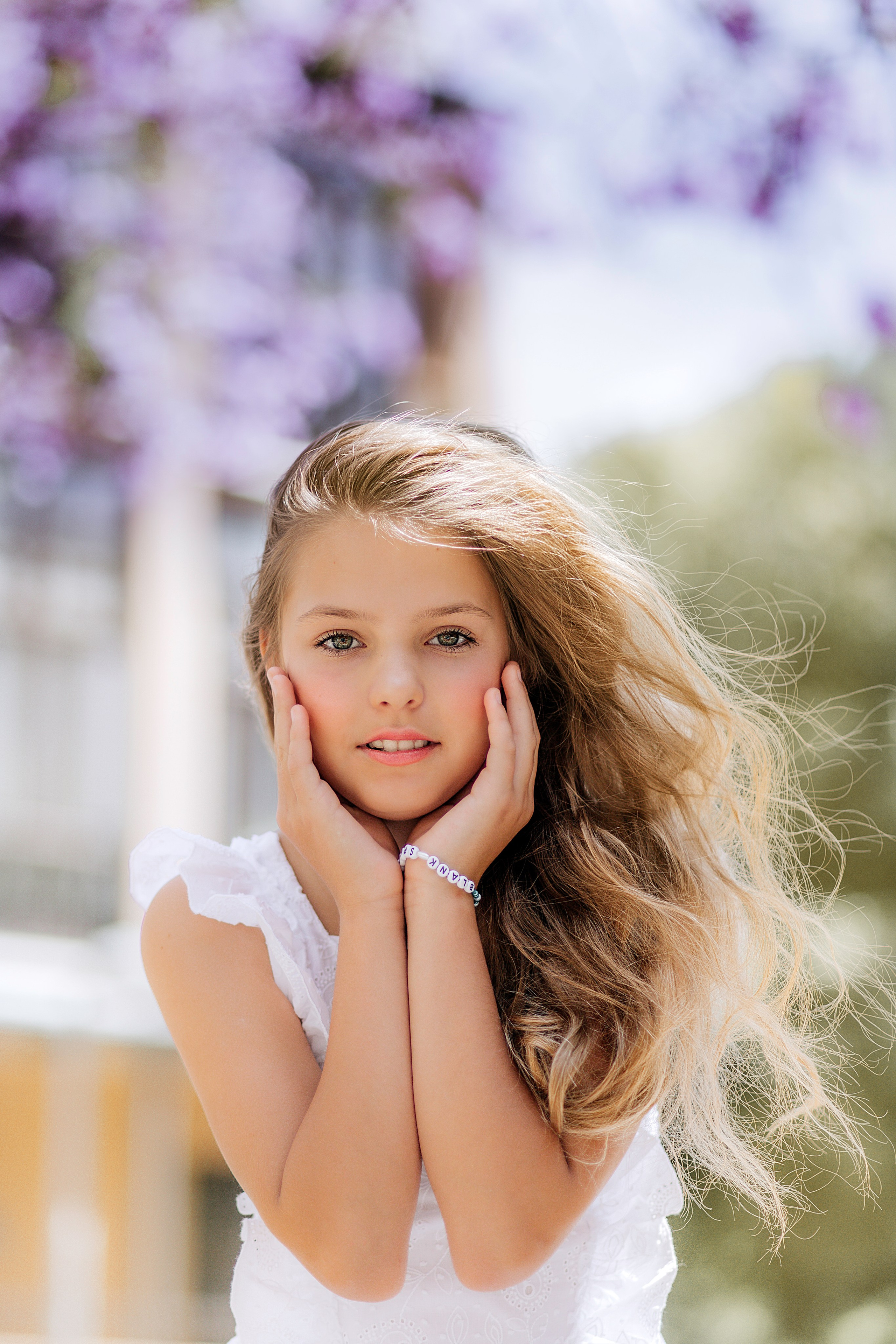 Причудливый уличный портрет молодой девушки с длинными светлыми волосами в белом платье, позирующей с руками на щеках под цветущими фиолетовыми деревьями жакаранды в Валенсии, Испания