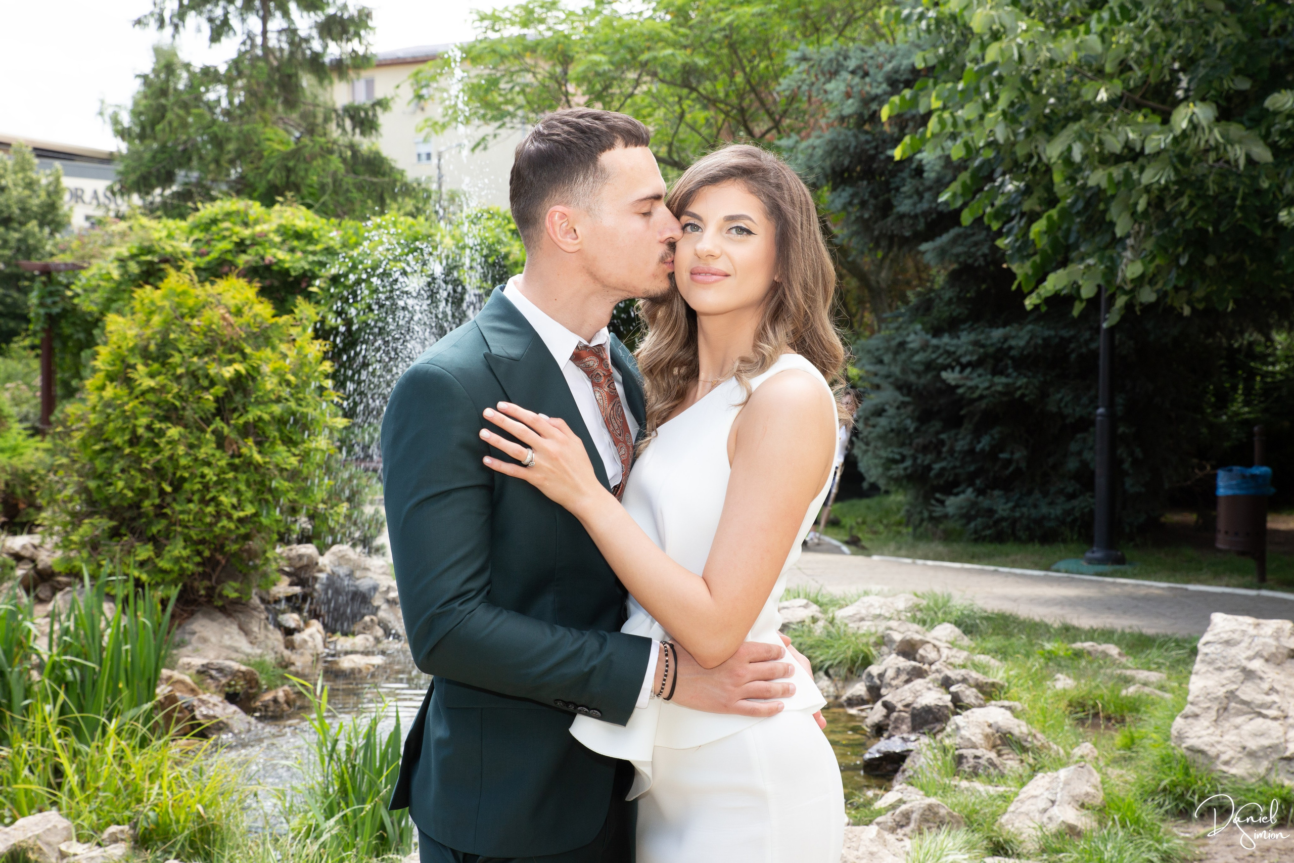 Man in suit kissing woman in white dress on cheek by garden fountain.