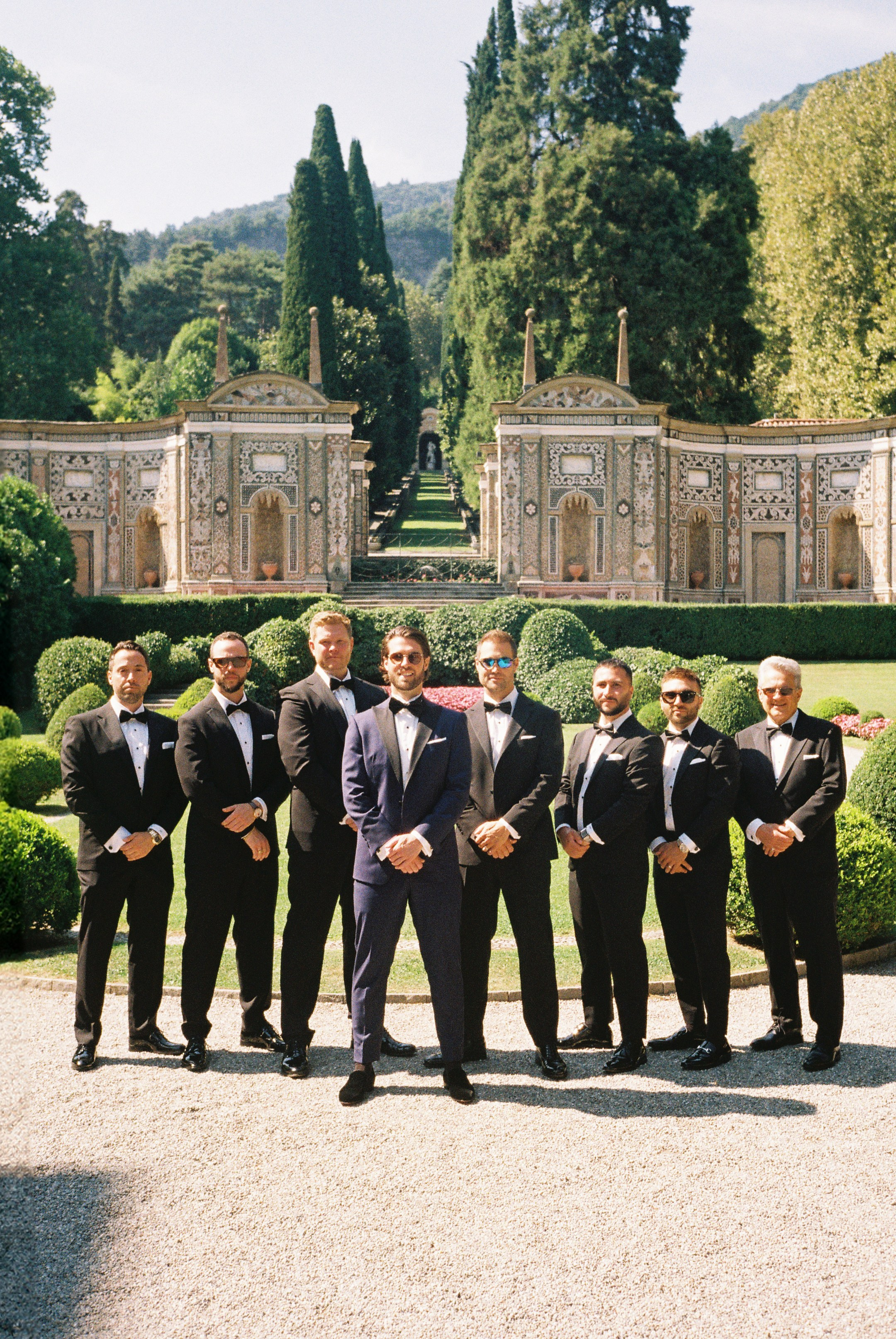 Groom and groomsmen in tuxedos posing in a formal garden with elegant stone architecture.