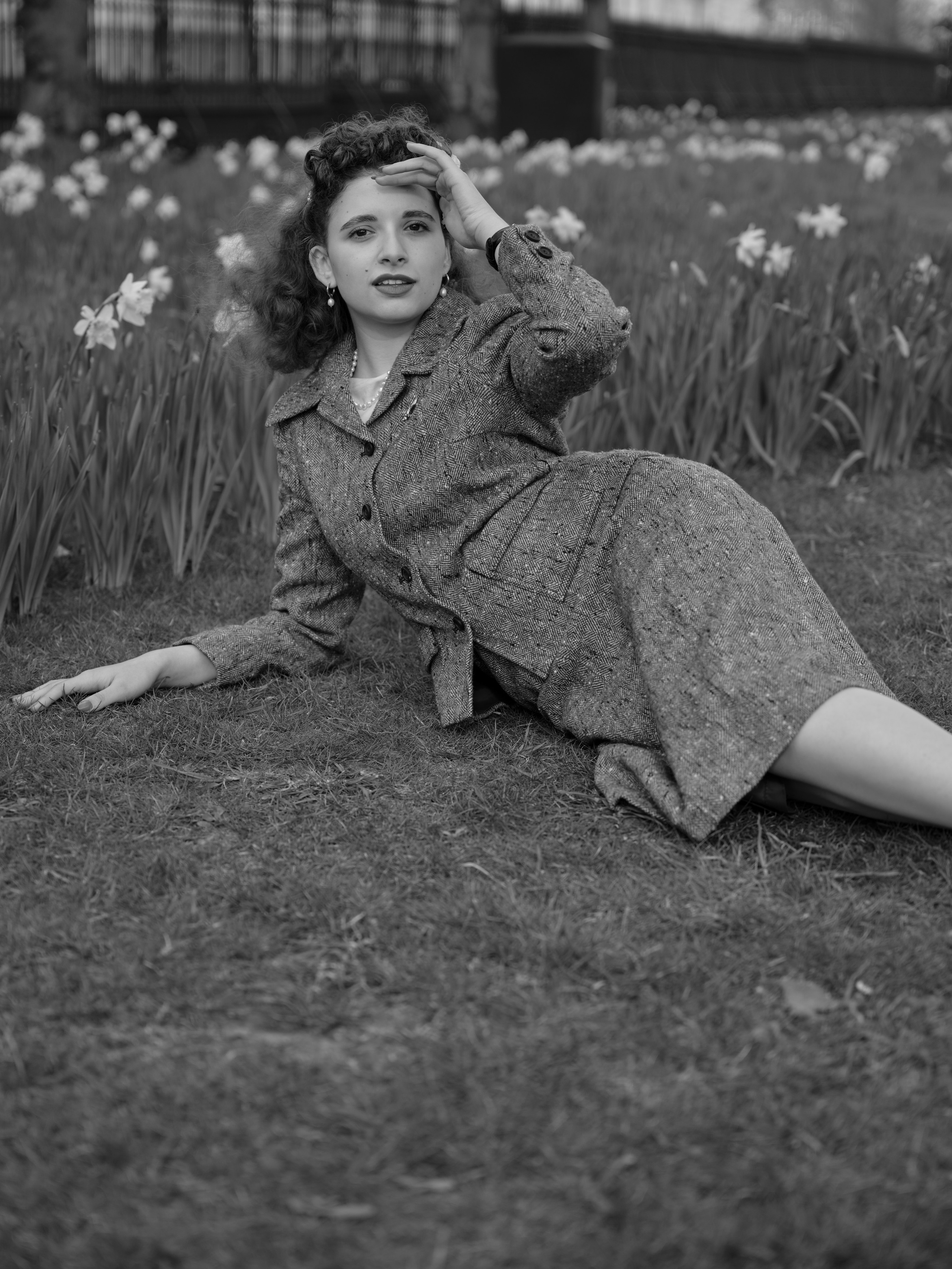 “Black-and-white full-length portrait of a woman in a vintage-inspired tweed suit reclining on the grass among rows of daffodils.”