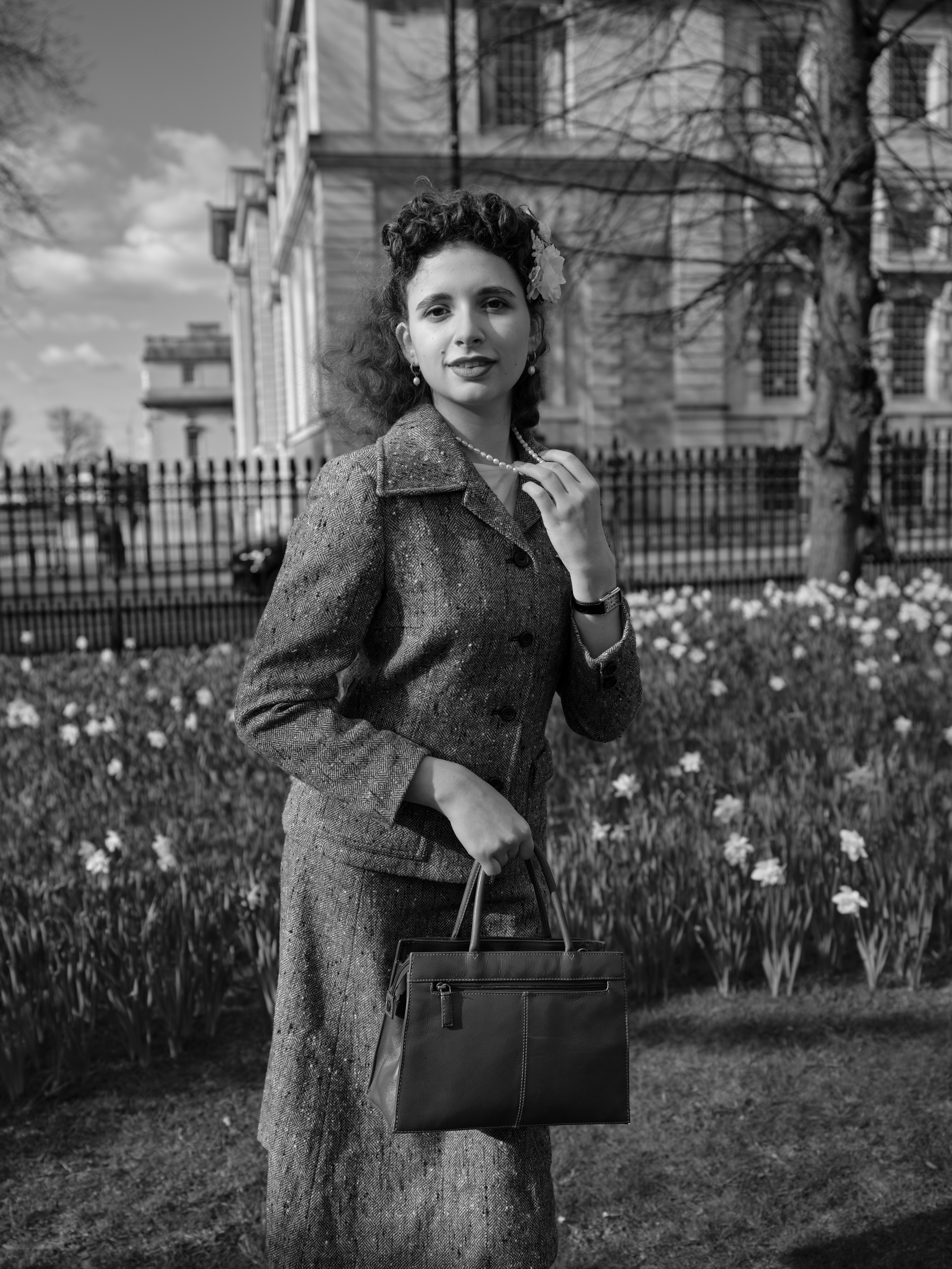 “Black-and-white portrait of a woman in a vintage-inspired tweed suit holding a structured handbag while standing among spring daffodils.”