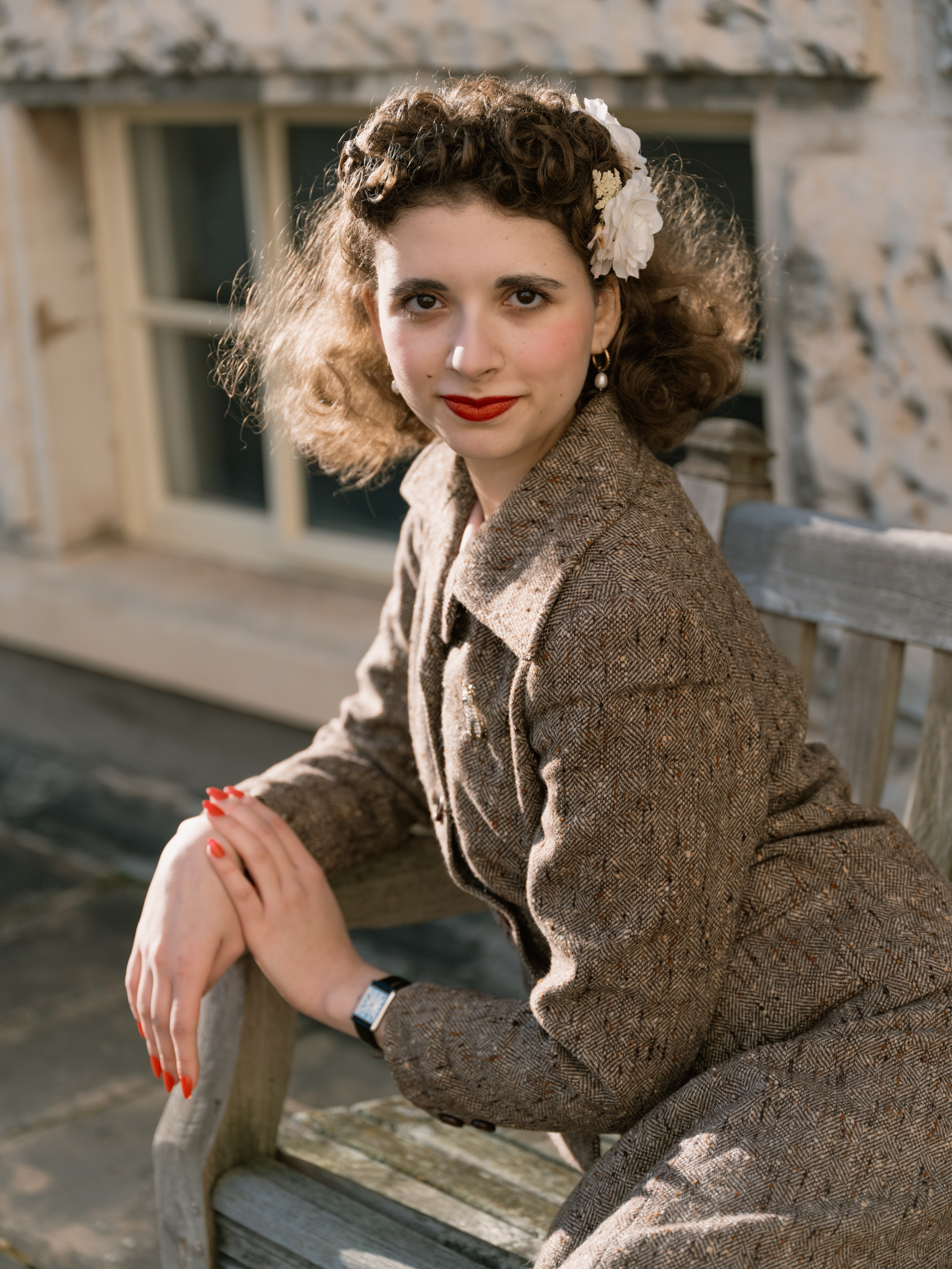 “Colour close portrait of a smiling woman with vintage-styled curls, white flowers and red lipstick, leaning forward with her hands beneath her chin.”