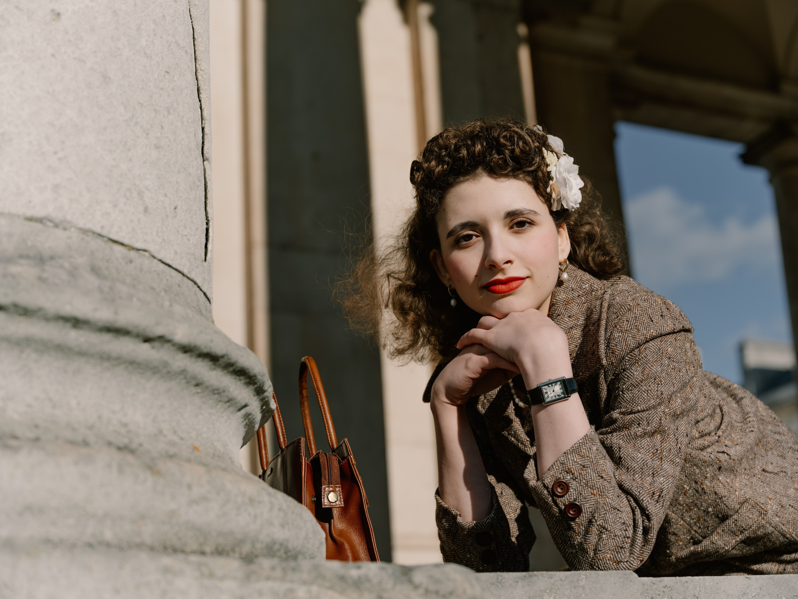 “Close colour portrait of a woman with curled vintage-styled hair and white flowers, resting her chin on her hands beside a brown leather handbag.”