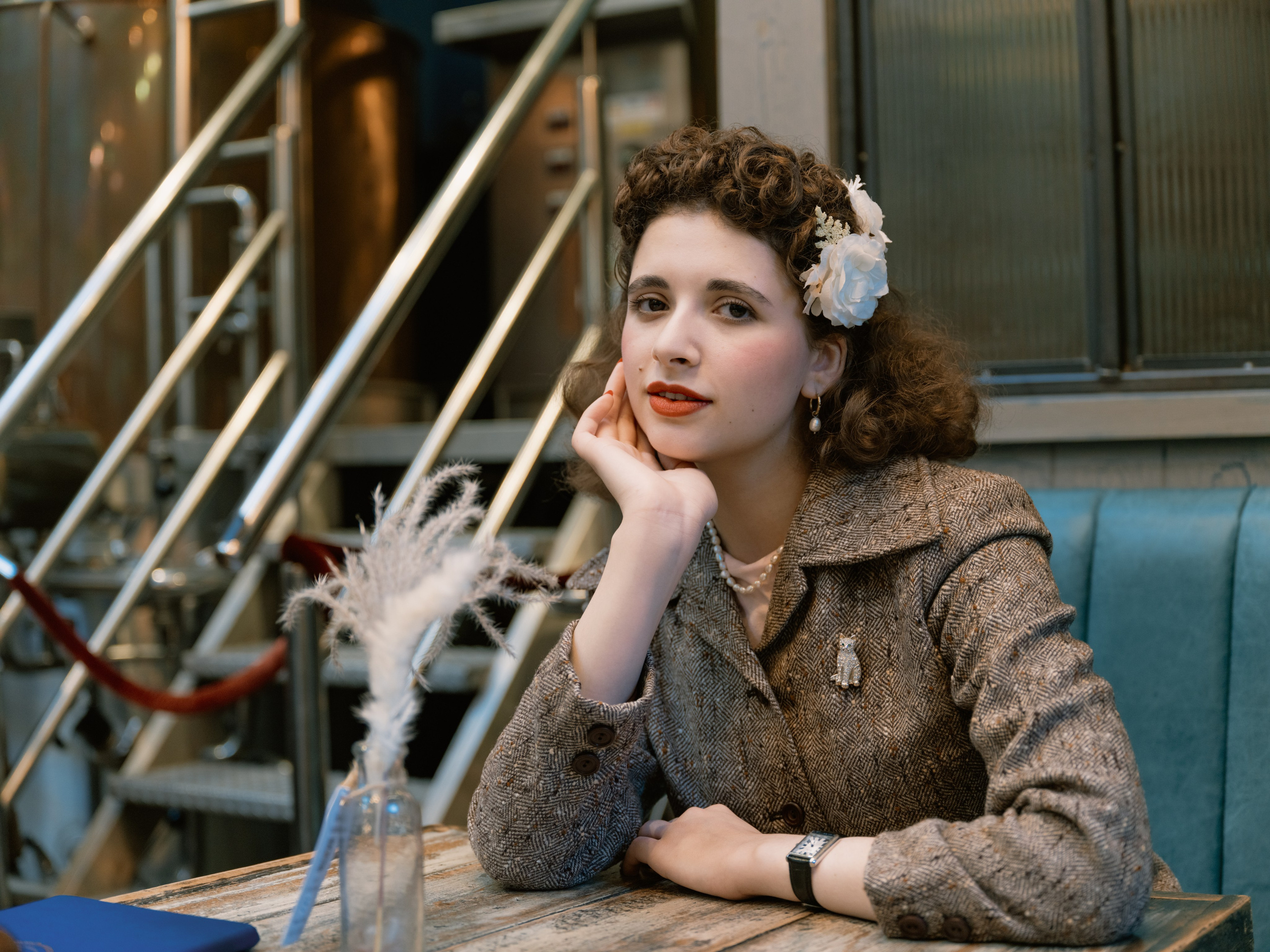 “Close colour portrait of a smiling dark-haired woman with vintage curls, white flowers and red lipstick, seated on a wooden bench in soft sunlight.”