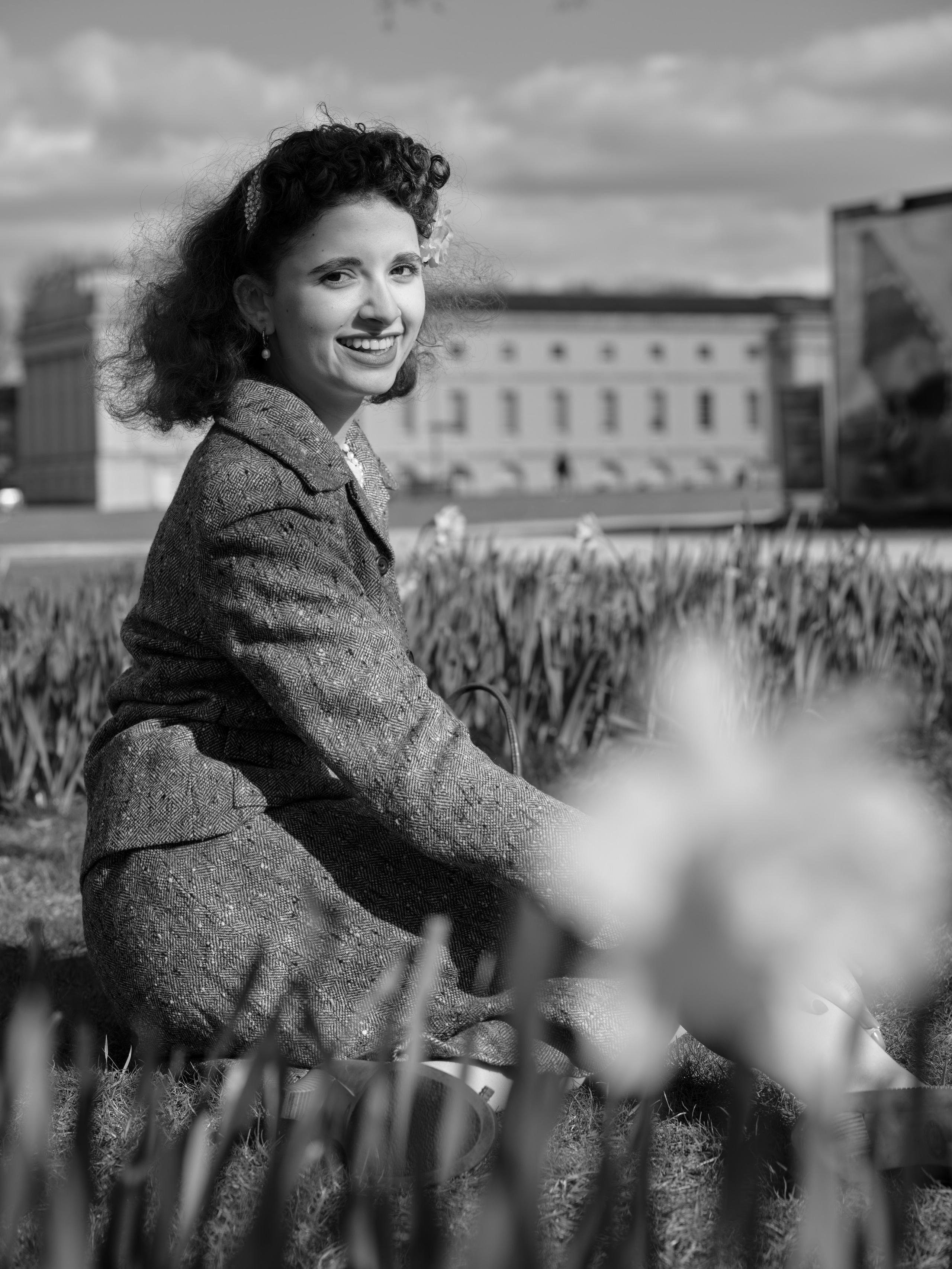 “Black-and-white close portrait of a smiling dark-haired woman in a tweed suit sitting among spring flowers, with classical architecture softly blurred behind her.”