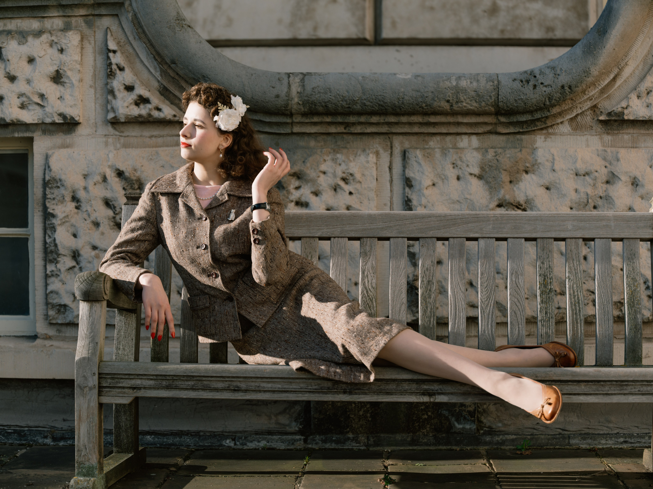 “Colour portrait of a dark-haired woman in a brown tweed suit seated on a wooden bench, looking directly toward the camera in warm sunlight.”