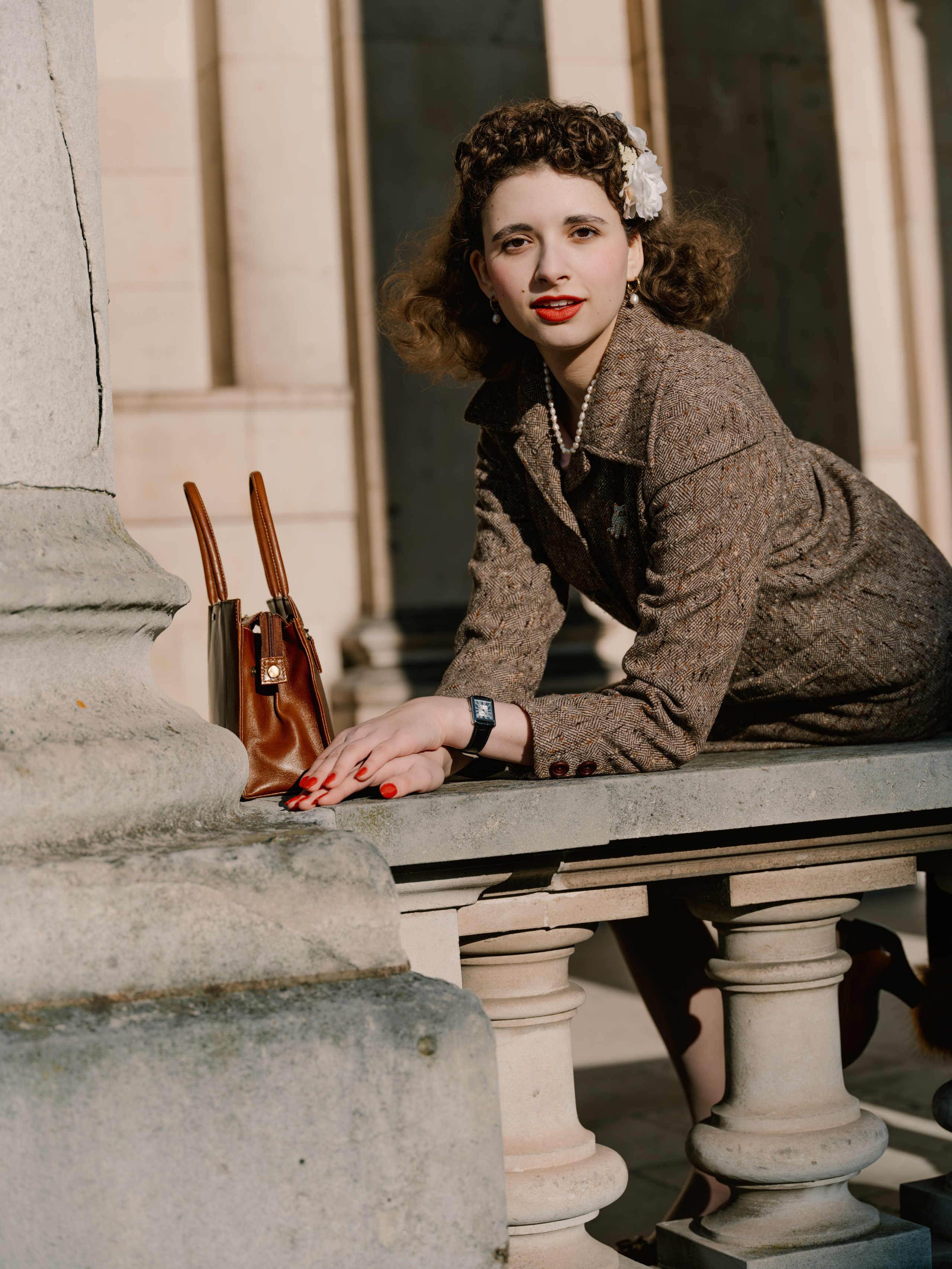 “Colour portrait of a woman in a brown tweed suit leaning on a stone balustrade beside a leather handbag, with red lipstick and nails adding bright accents.”