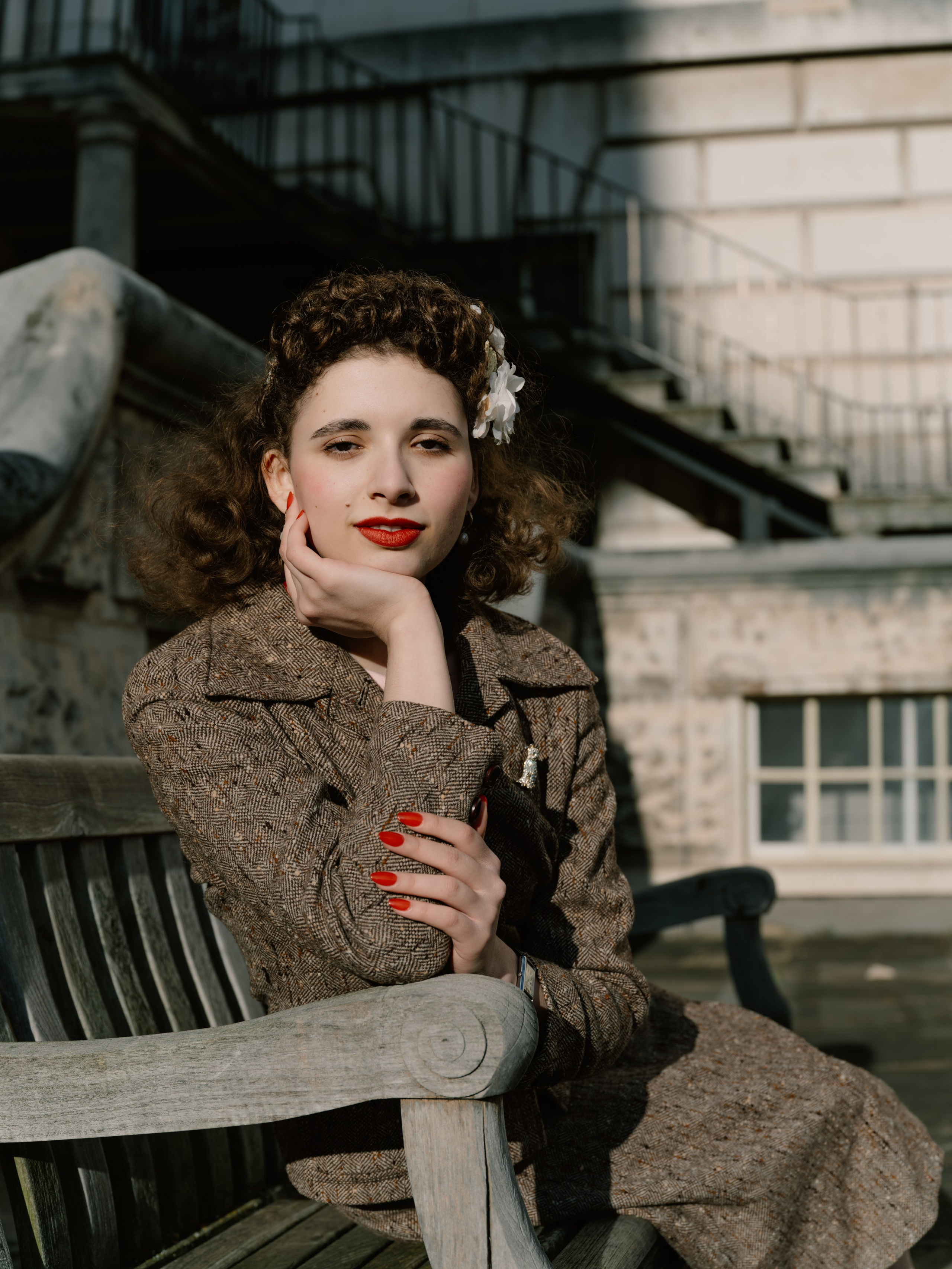 “Colour portrait of a woman in a brown tweed suit sitting on a wooden bench, resting her face in one hand against a sunlit architectural background.”