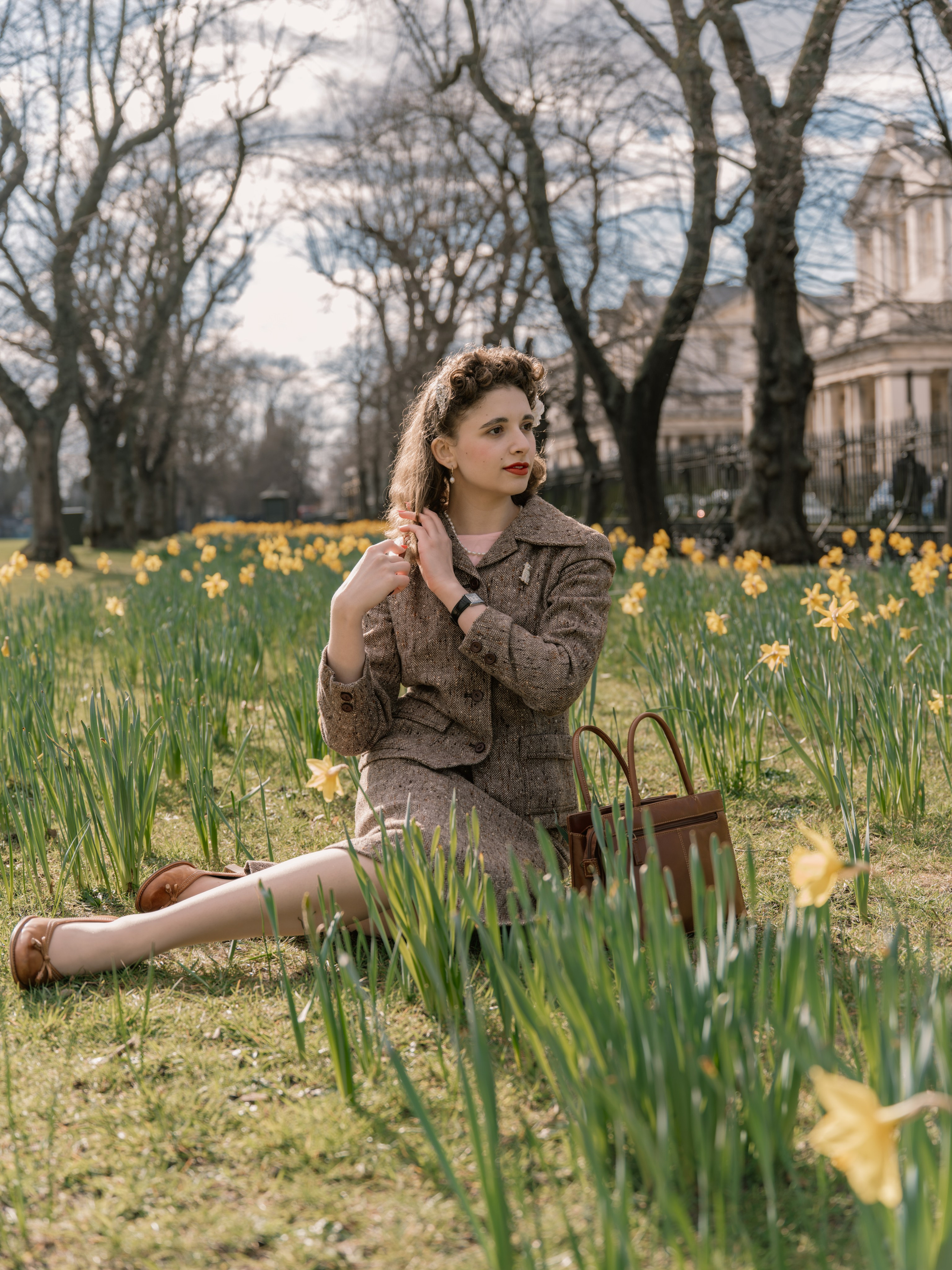 “Colour portrait of a woman in a vintage-inspired brown tweed suit sitting among yellow daffodils in a London garden, with historic architecture behind her.”