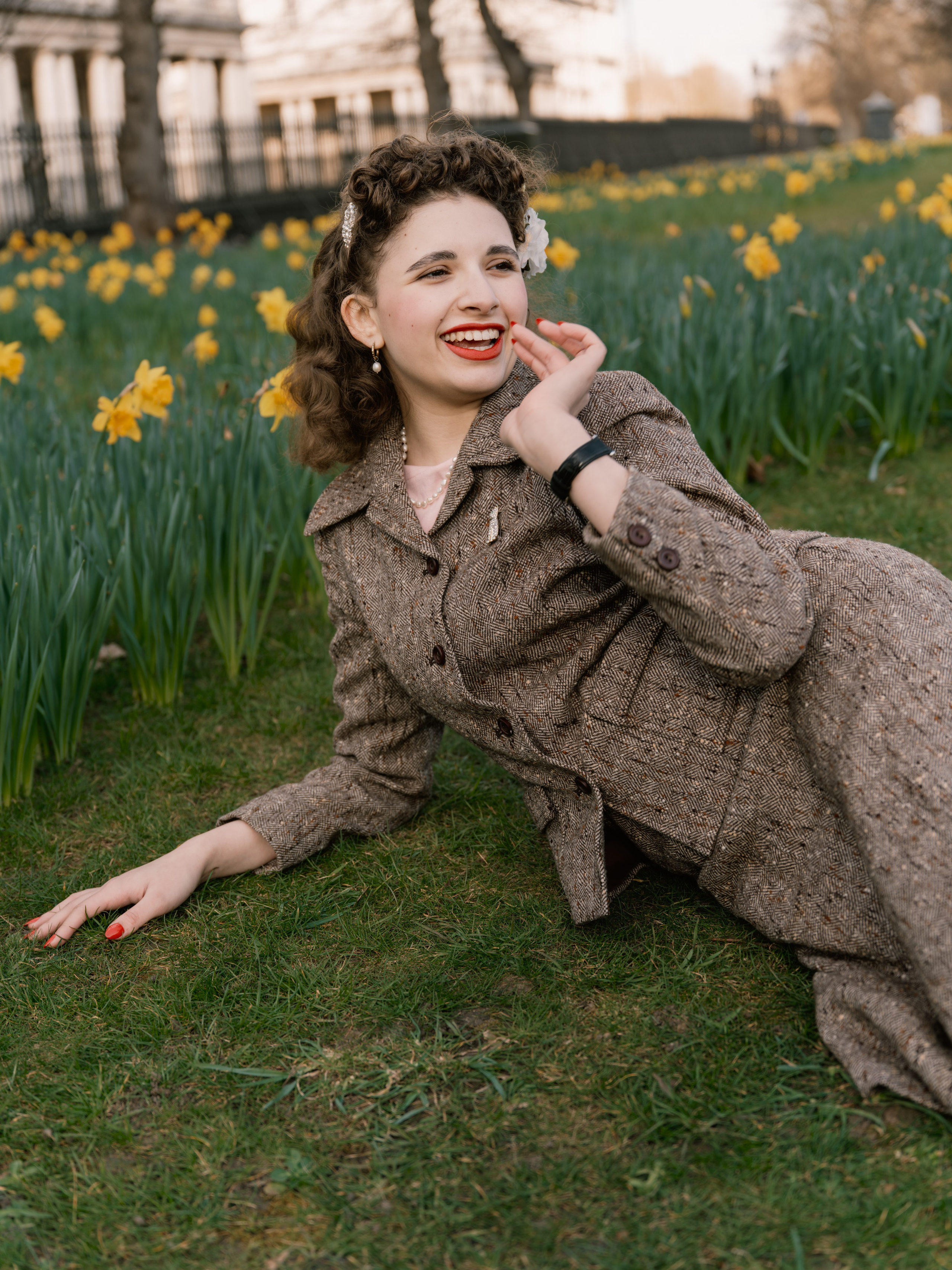“Colour close portrait of a laughing woman with vintage-styled hair, red lipstick and a brown tweed suit, surrounded by yellow daffodils.”